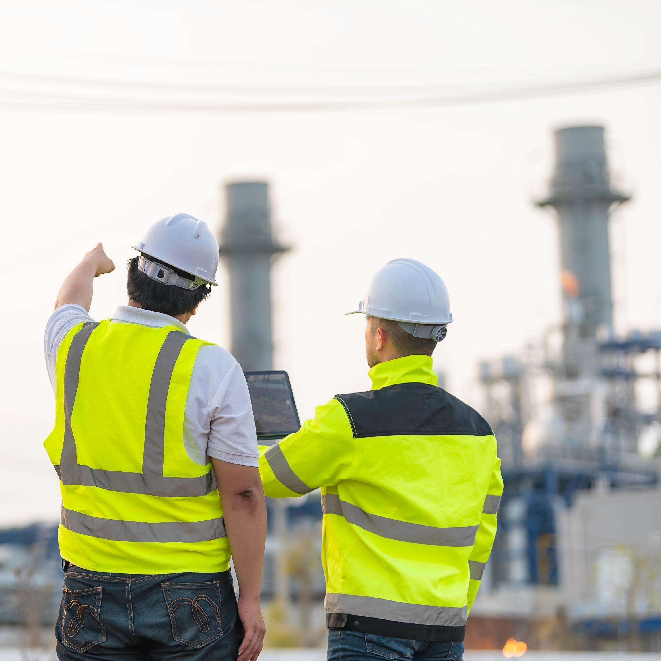 Two experts from behind in front of a refinery. One is pointing at something in the distance, the other is holding a laptop.