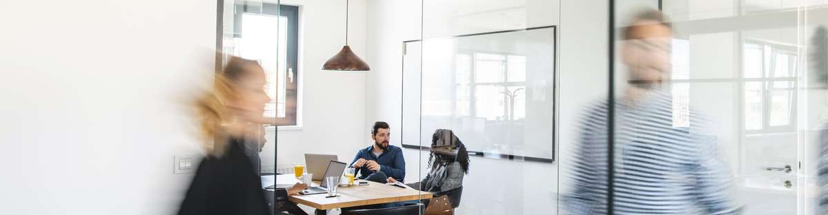 Three people are sitting in a meeting room made of glass and are talking while some people are walking by in front of the room.