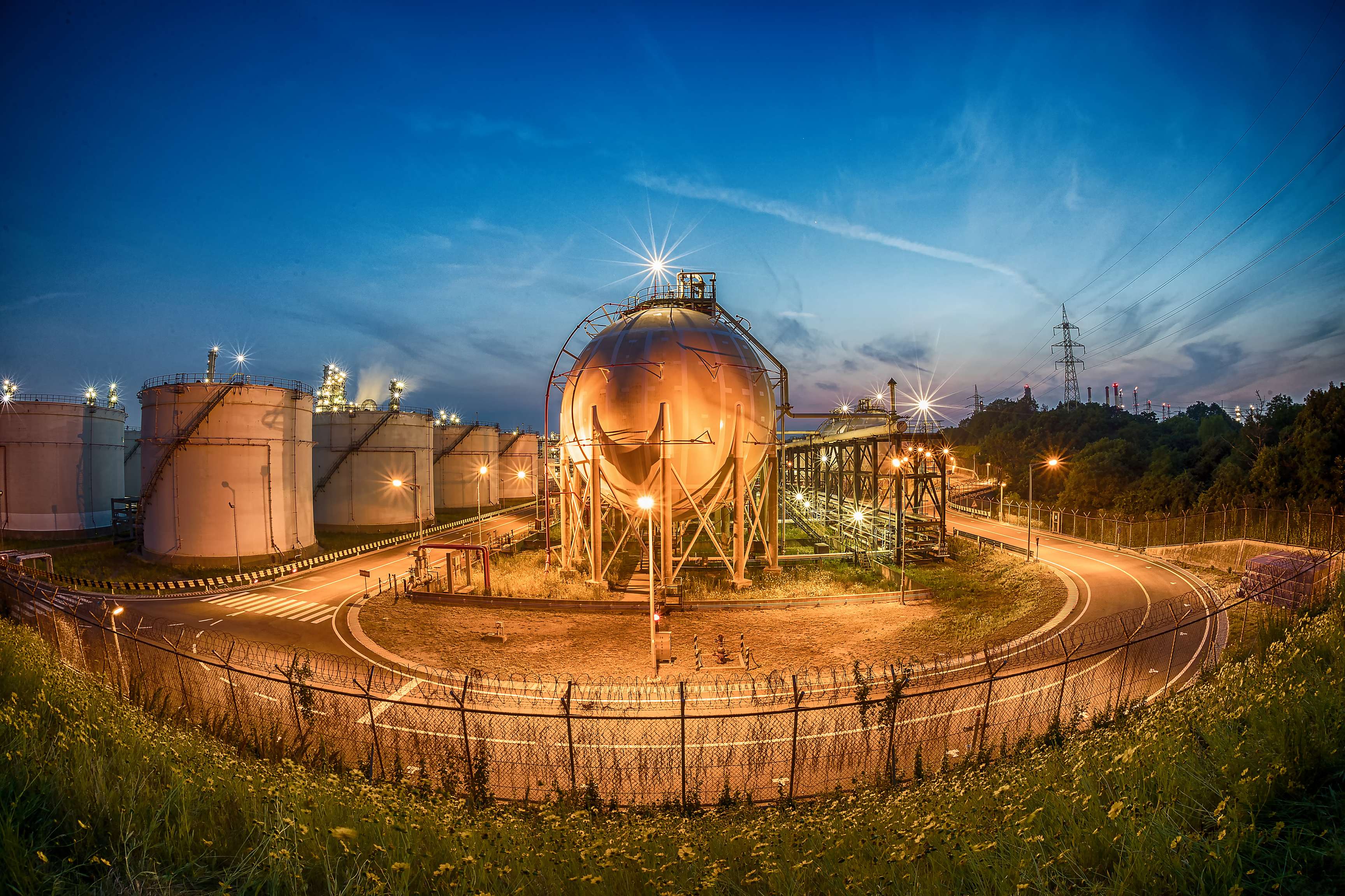 Pressure vessels at dusk next to a tank farm with city lights in the background.
