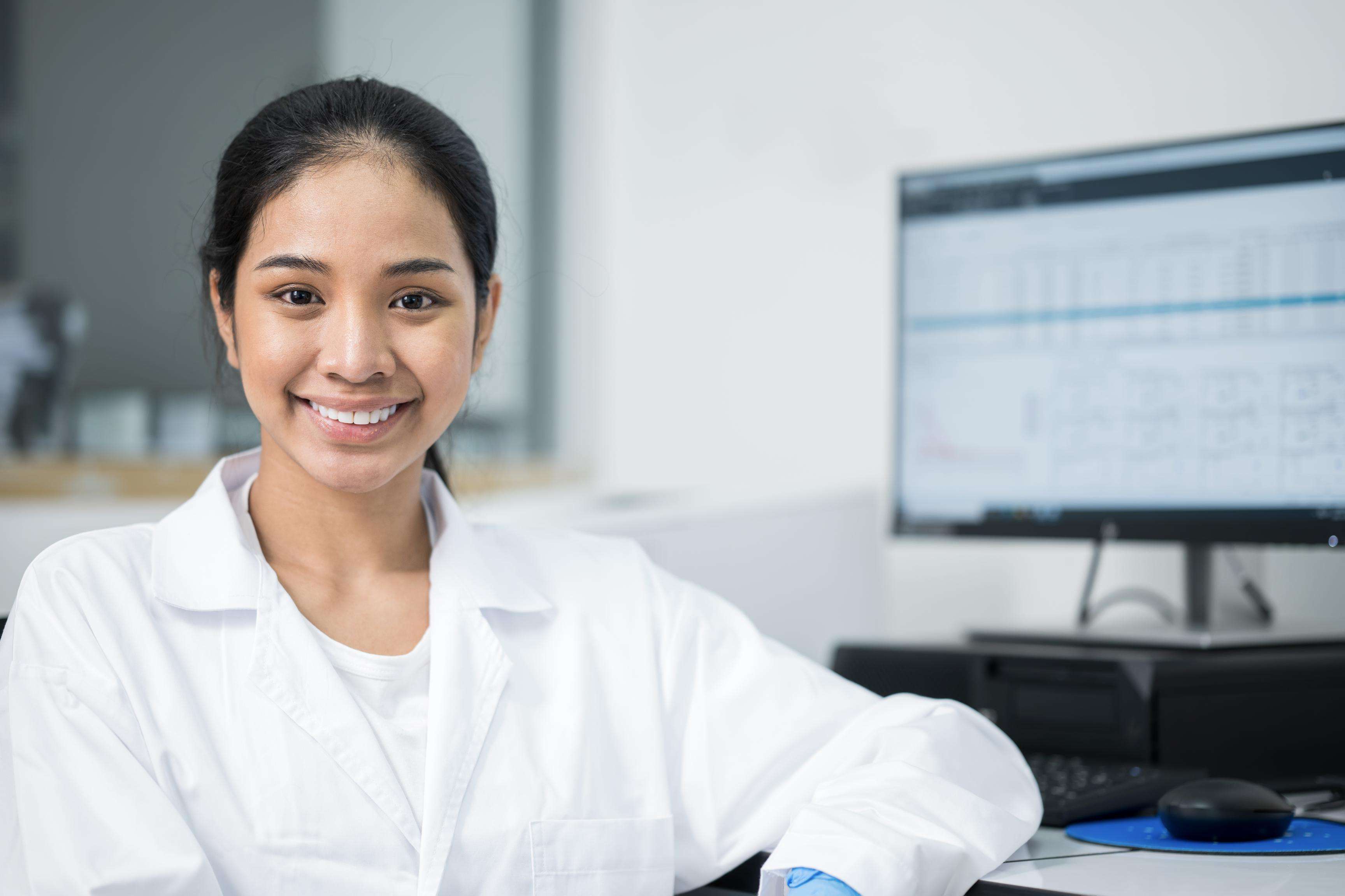 Young woman wearing white lab coat and sitting in front of screen.