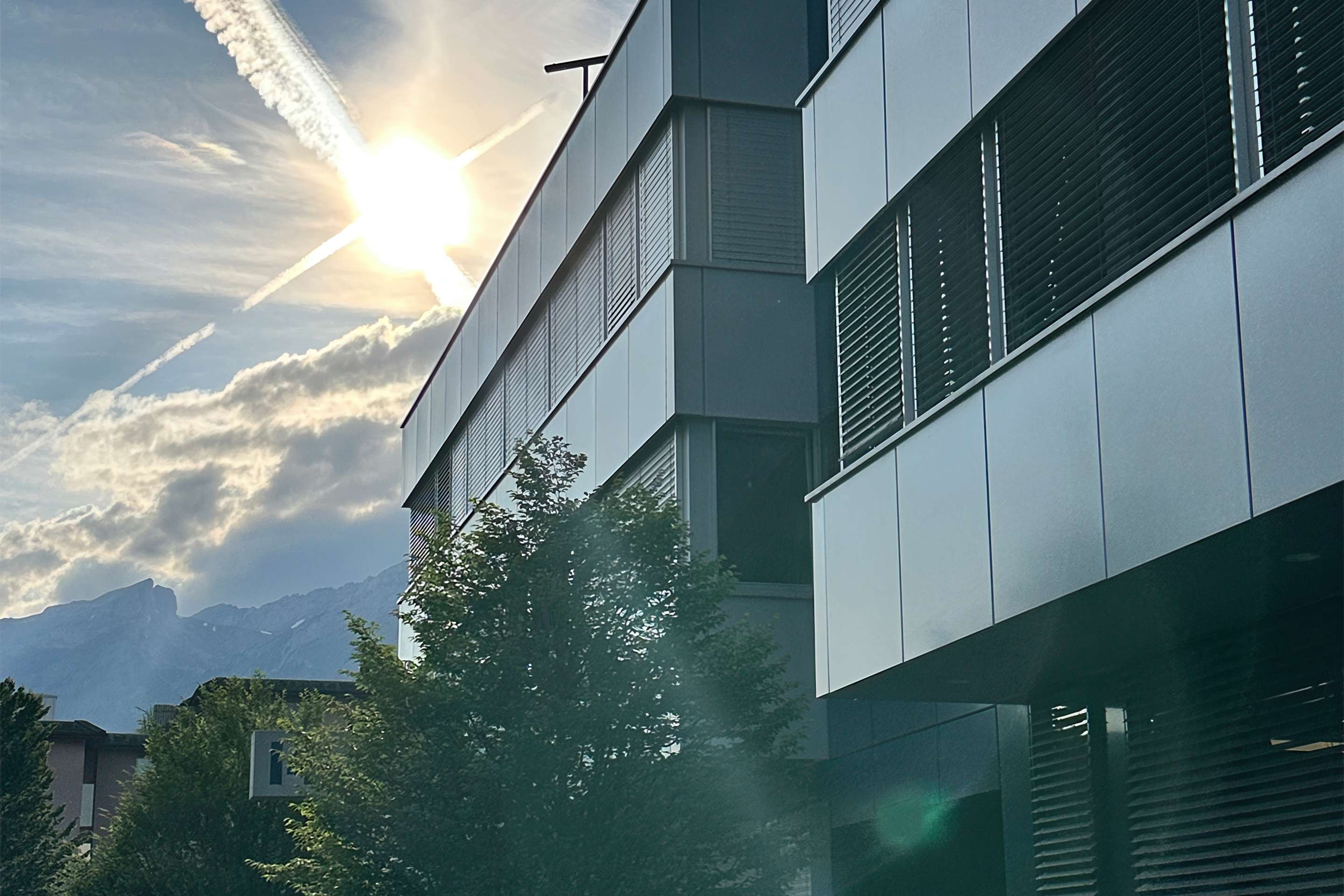 The photo shows the ROSEN Group headquarters in Switzerland, with its metal facade, backlit by the sun and surrounded by trees, with a mountain landscape in the background.
