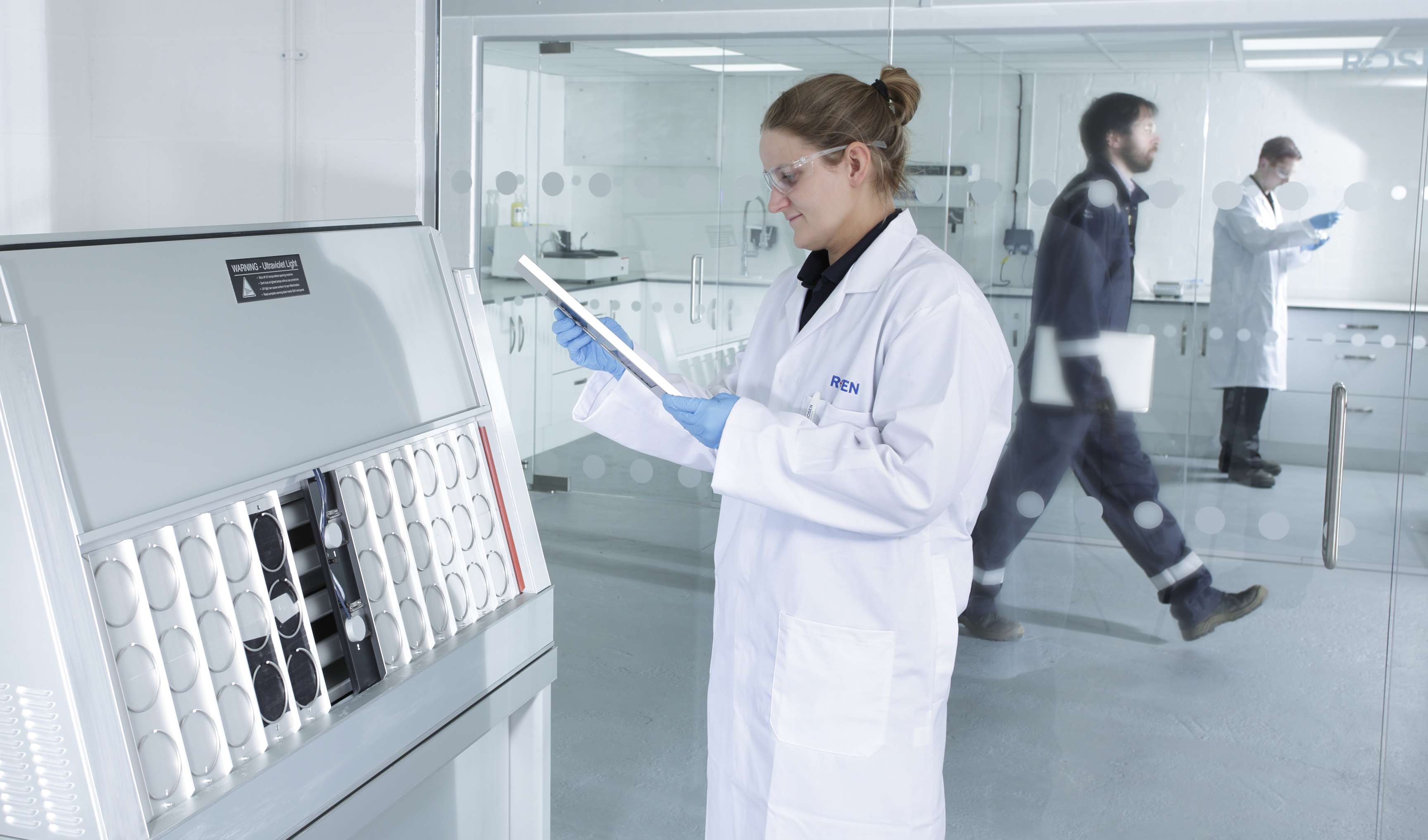 Employee stands in front of a machine in a lab wearing safety goggles and looks at a clipboard.