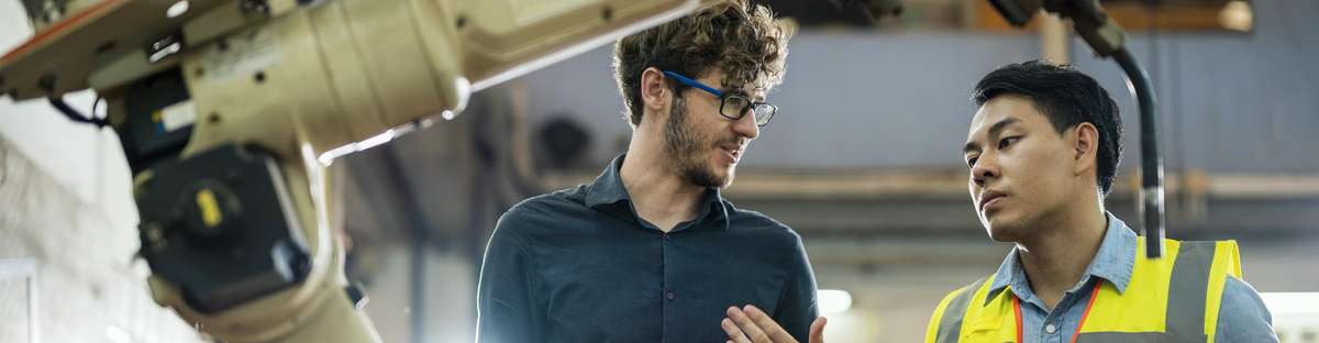 Two men talking to each other and standing next to a robot arm.