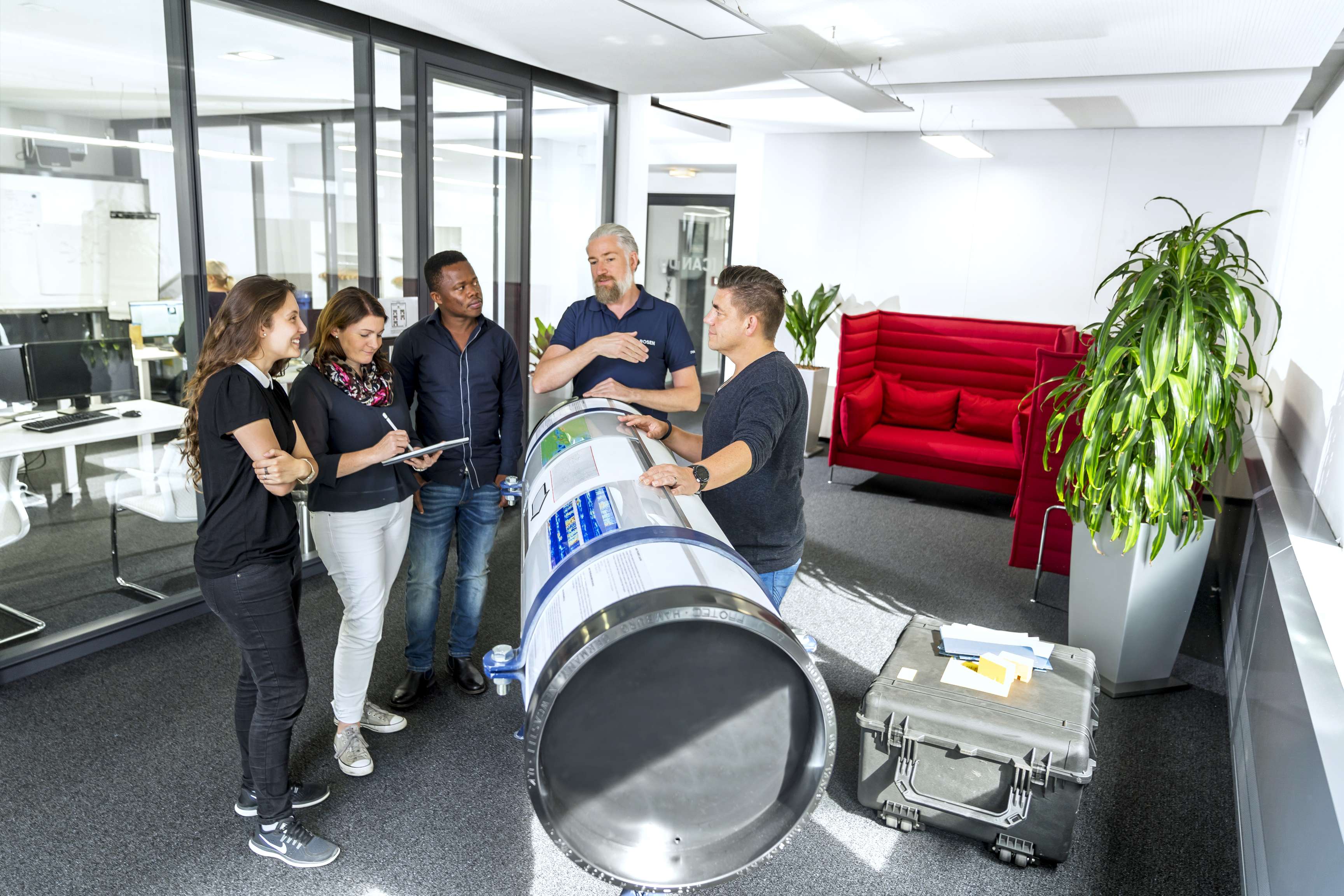 Five people standing around a labeled pipeline model explain something or take notes.