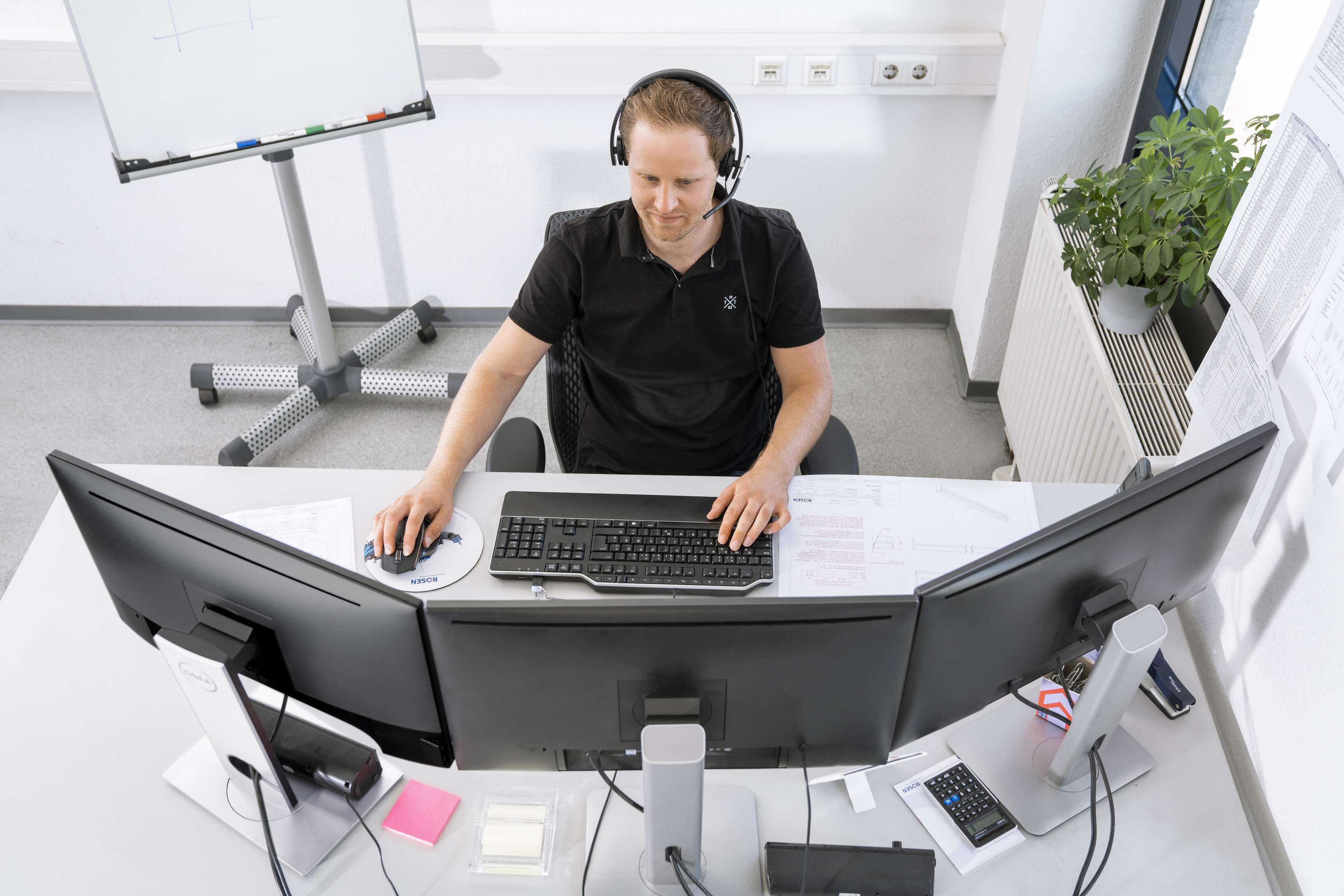 Ein ROSEN Mitarbeiter sitzt an einem Schreibtisch mit drei Monitoren und trägt ein Headset. Die Situation ist frontal von vorne fotografiert und soll eine Arbeitssituation in der Arbeitsvorbereitung abbilden.