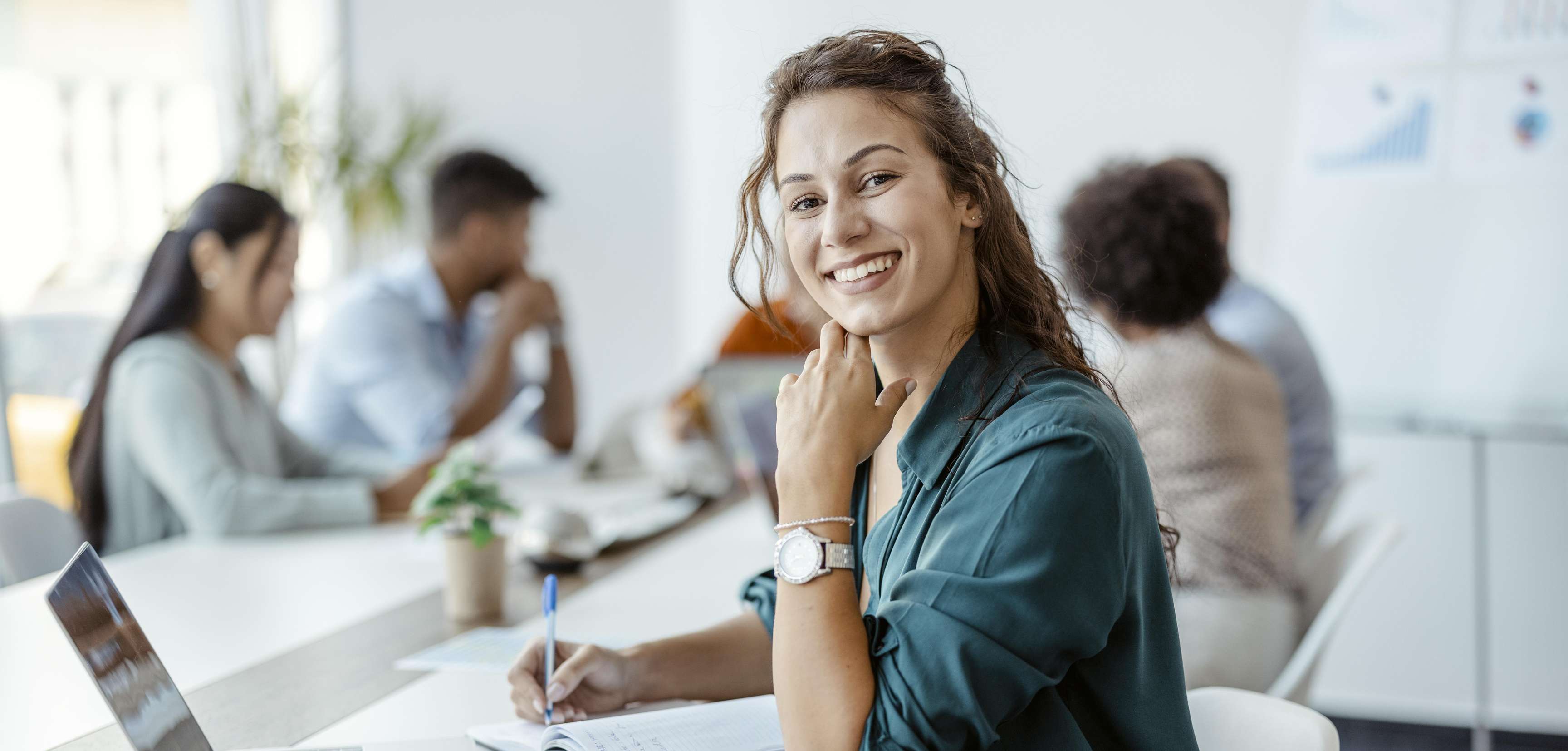 Woman sitting in the office in front of her laptop, taking notes and smiling at the camera, while her colleagues can be seen in the background.