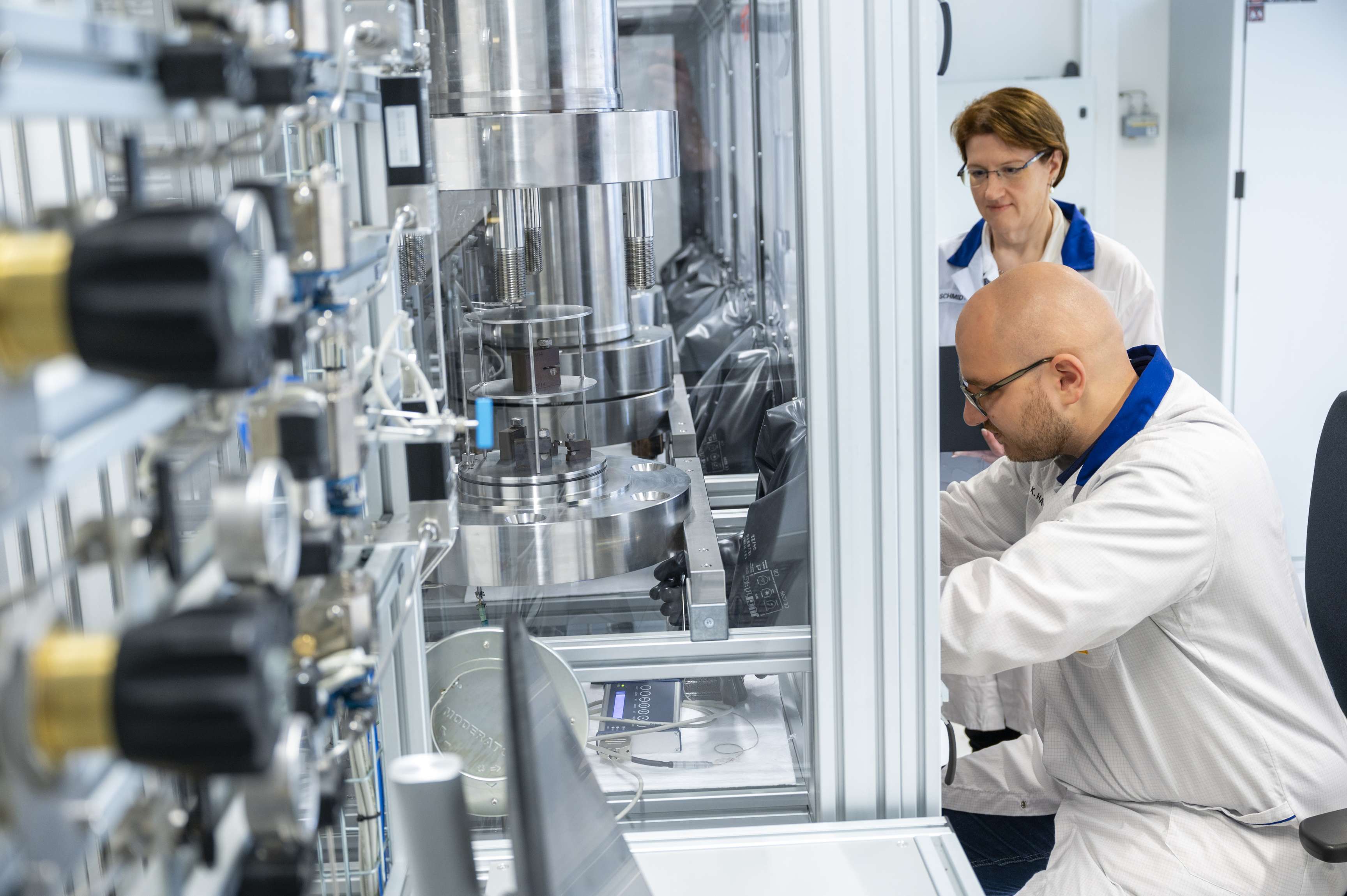 Employee works in a booth in a laboratory while his colleague stands next to him and watches.