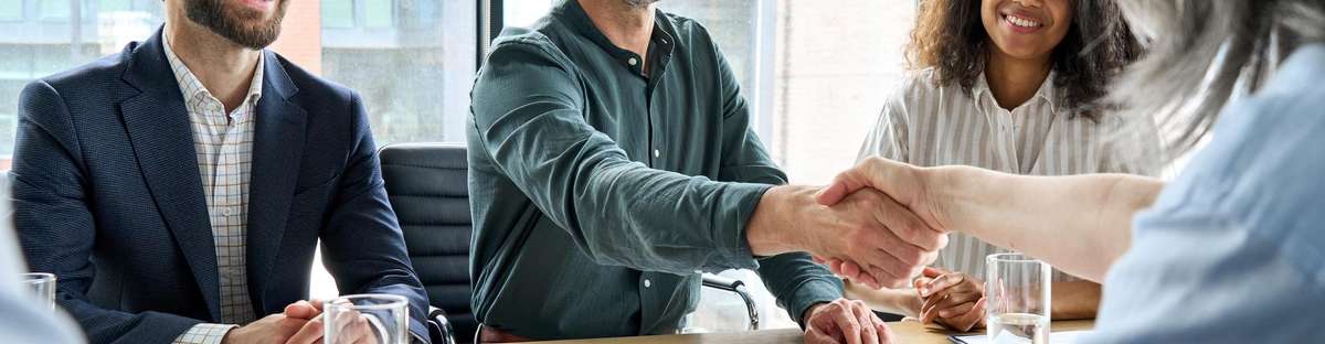 Several people in business attire are sitting around a table with notes and water glasses in front of them, two of them shaking hands.