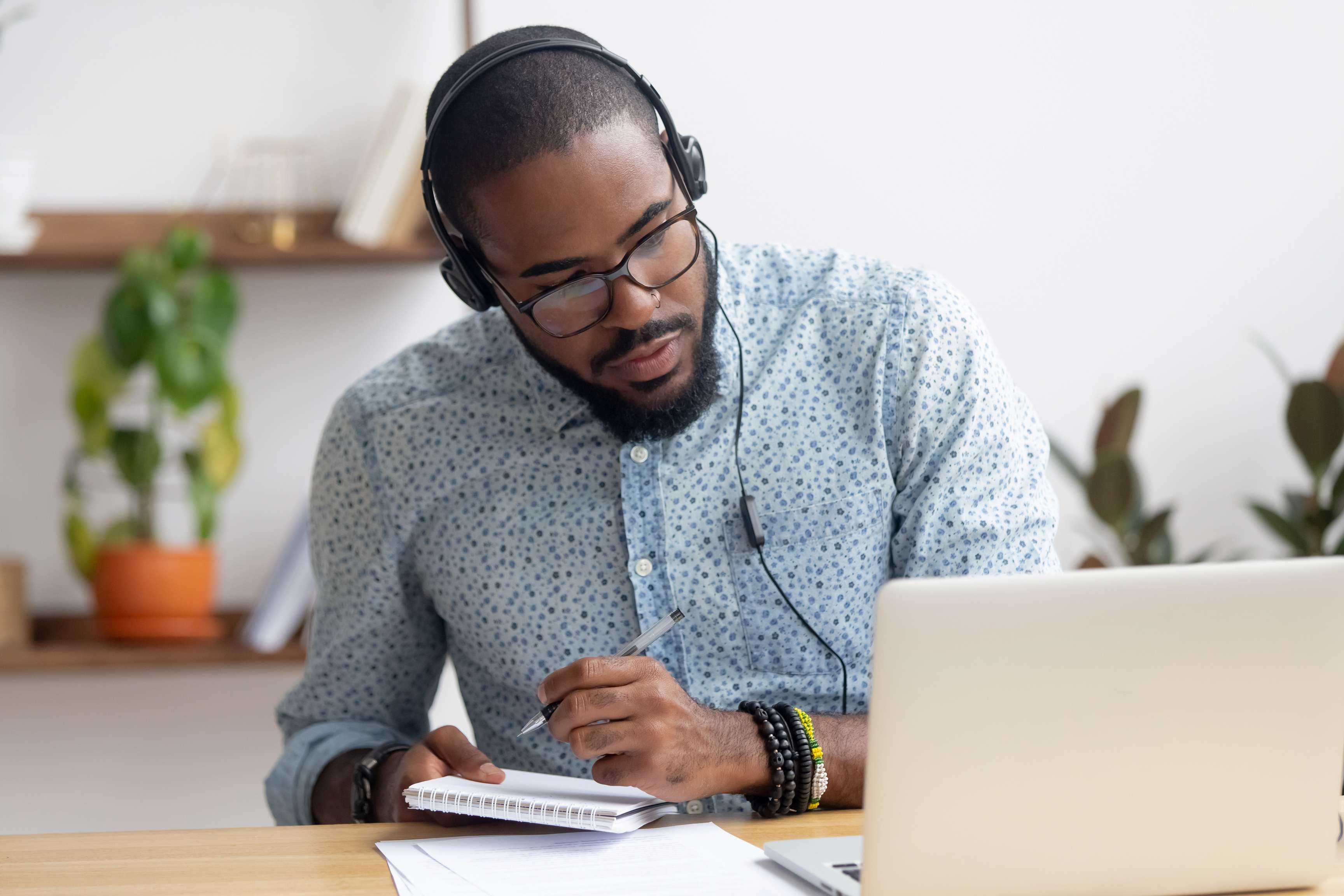 Man with headset and glasses sitting in front of laptop and taking notes.