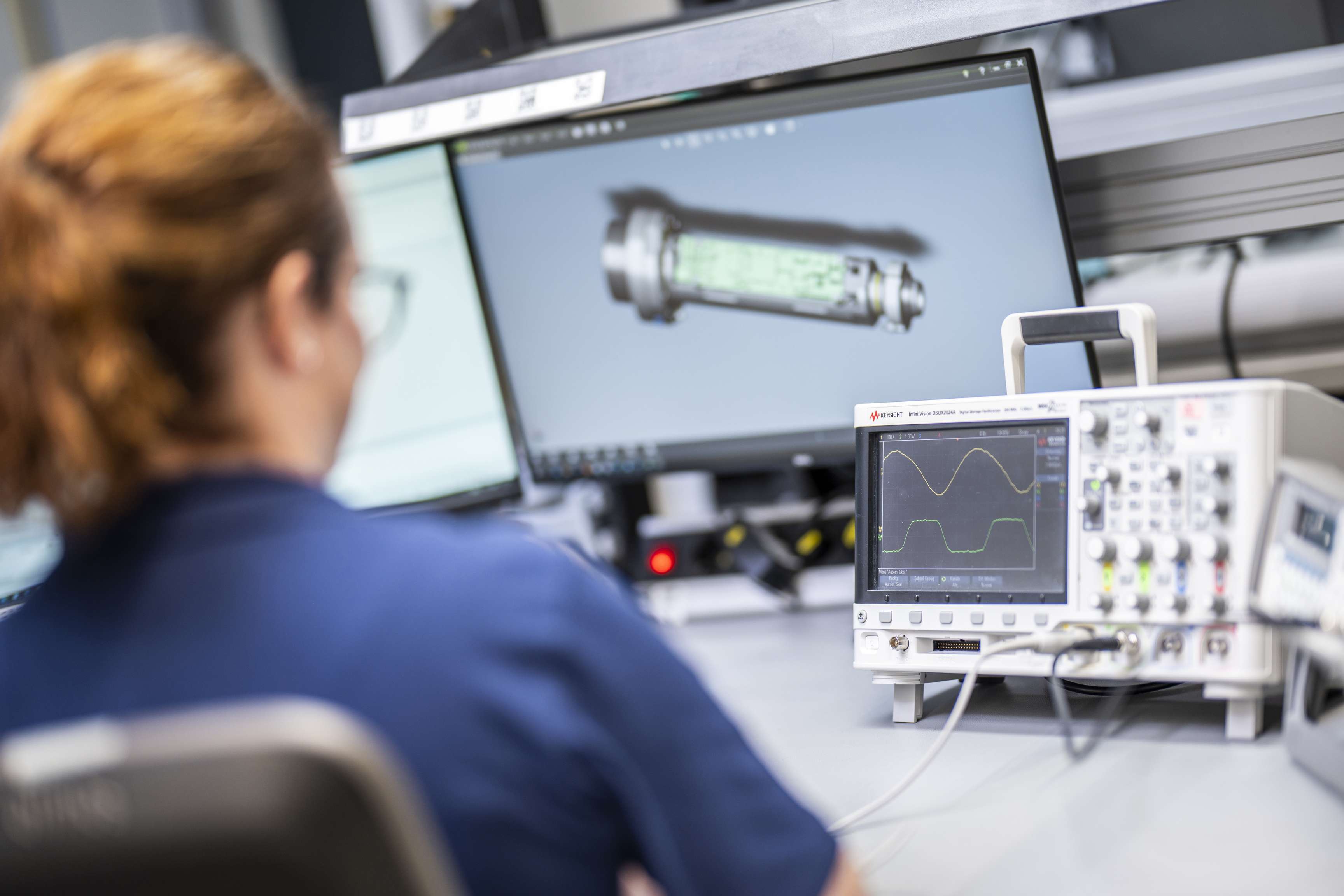 Eine Frau sitzt vor einem PC Bildschirm und einem Messgerät und schaut sich die gemessenen Wert an