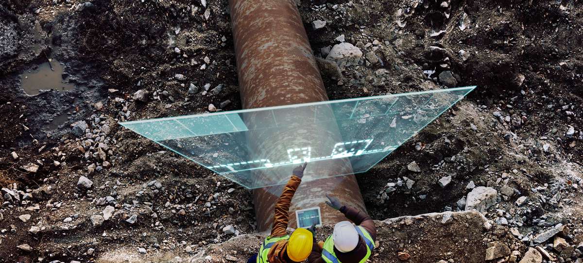 two workers standing next to a rusty, dug out pipeline looking at a hologram of a corrosion growth analysis