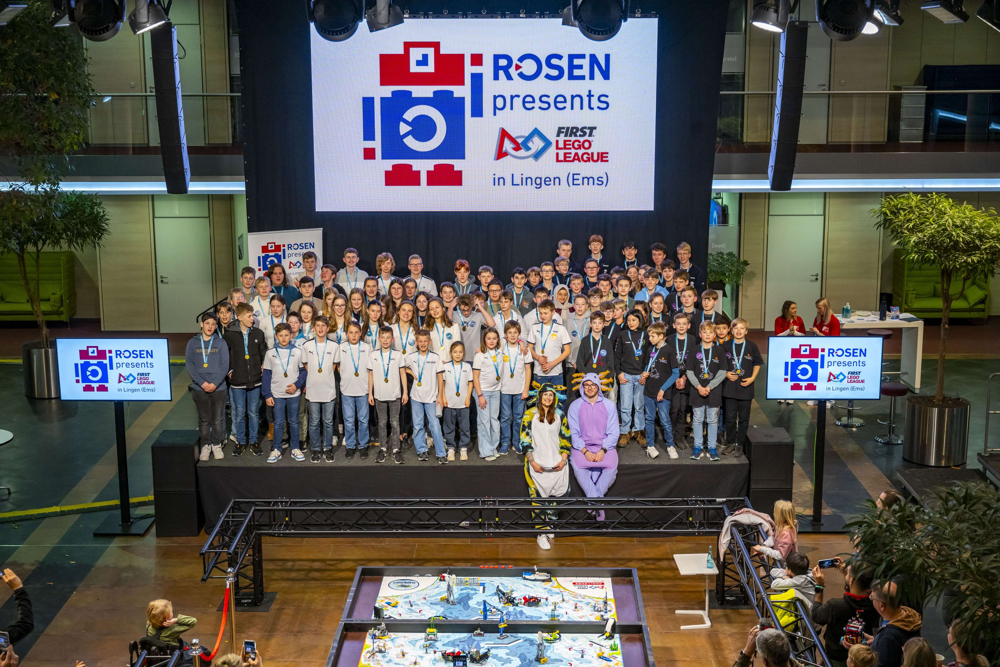 The participants of the First LEGO League are standing on a stage with the moderators. The participants are all wearing medals around their necks.