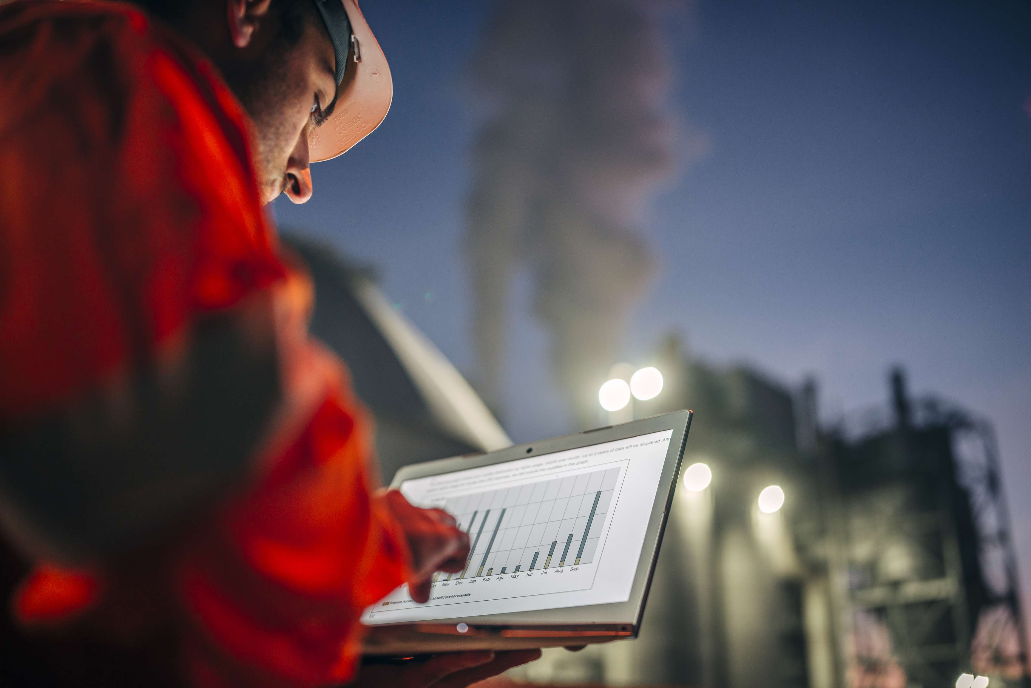 Expert in front from behind in orange jumpsuit looking at graphics on his laptop in front of a refinery at night.