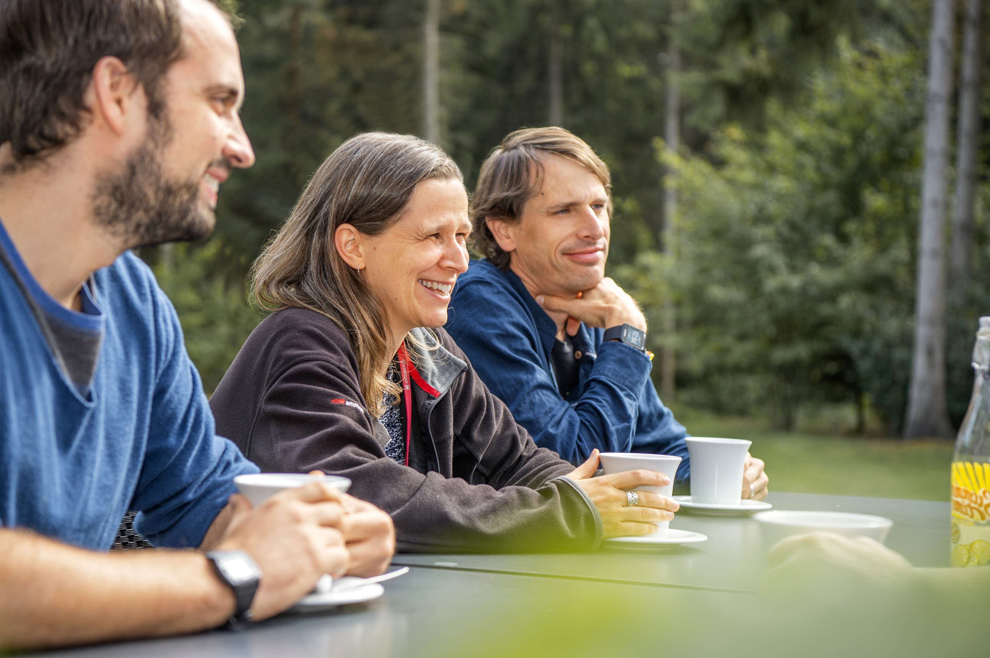 Three colleagues sit outside at a table holding mugs in their hands and trees in the background.
