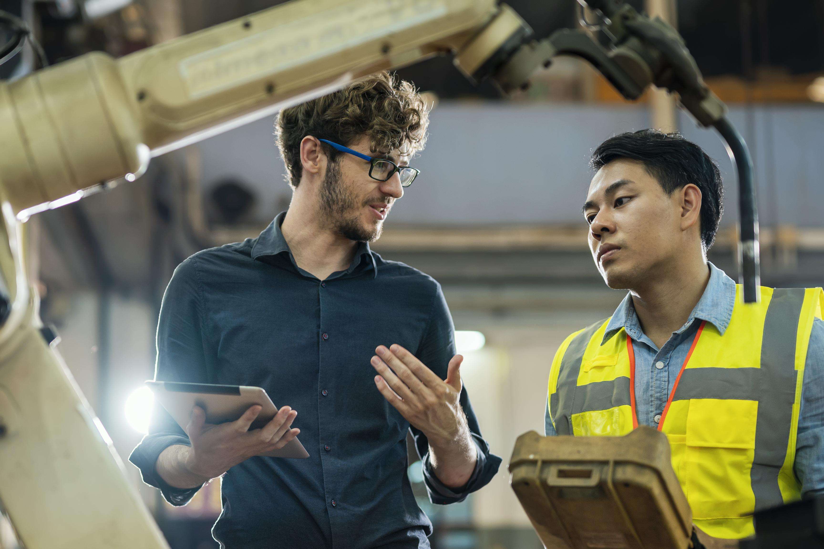 Two men talking to each other and standing next to a robot arm.