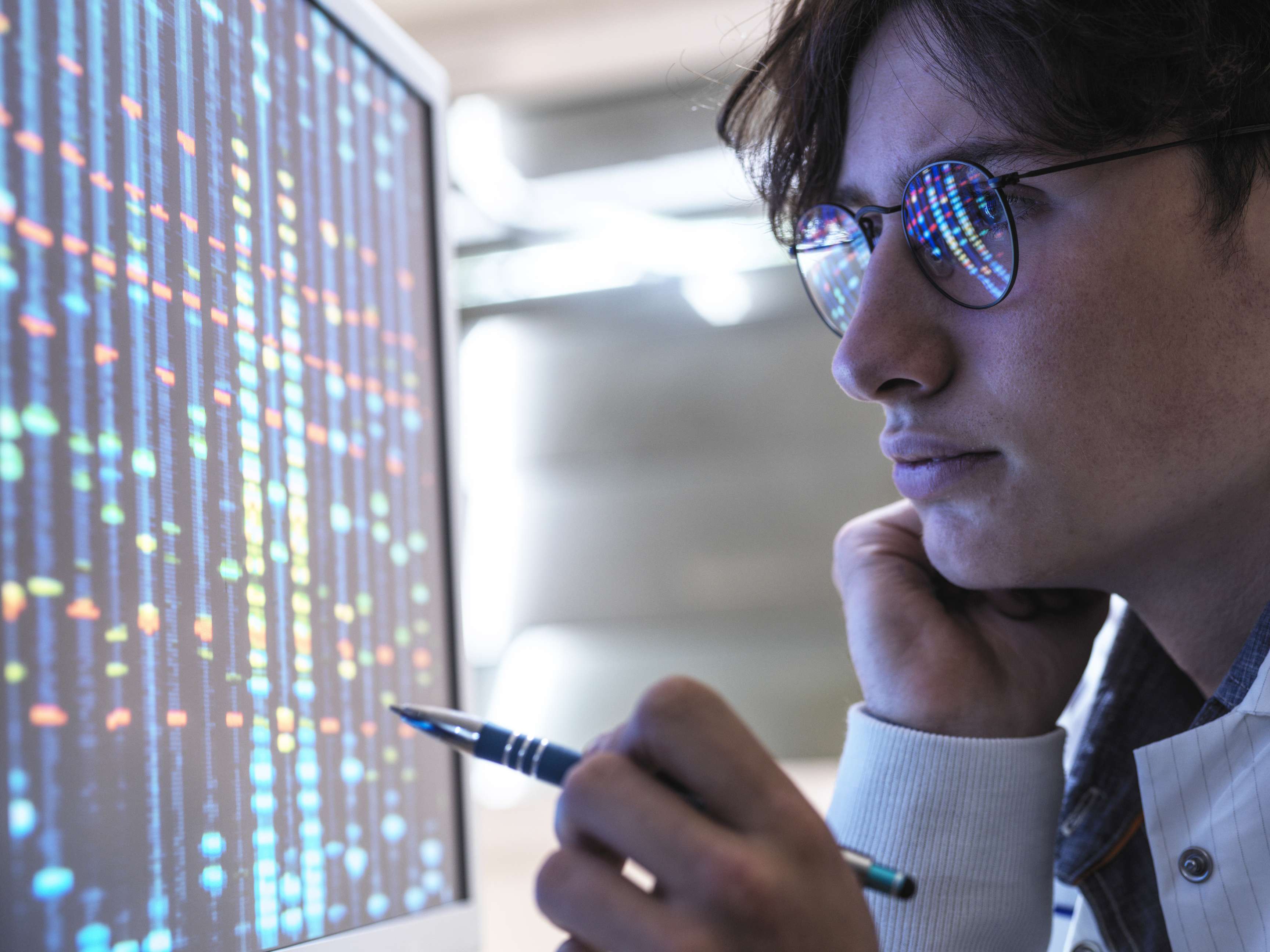 A man wearing glasses in front of a screen, holding a ballpoint pen while analyzing data.