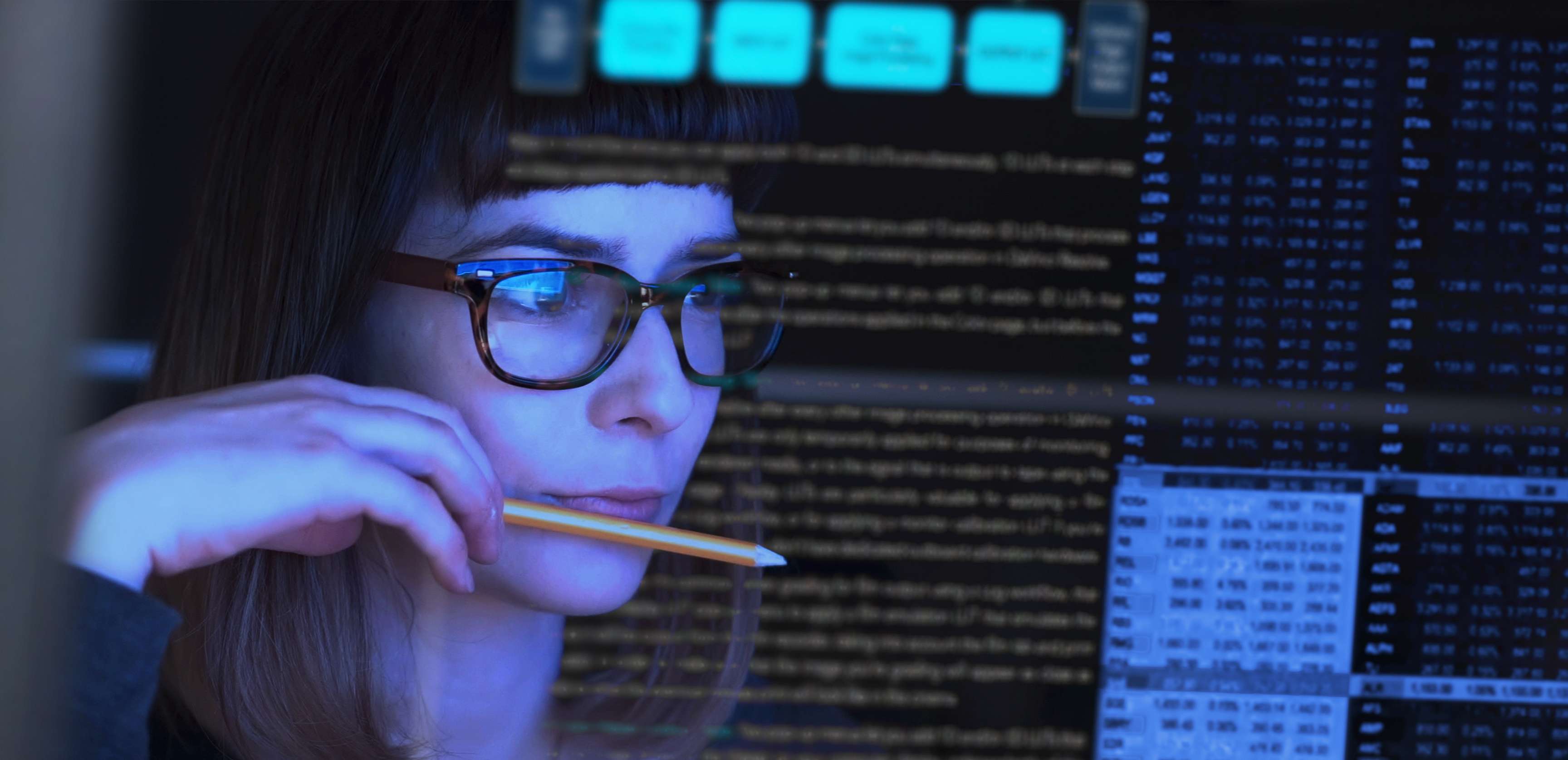 Woman wearing glasses and holding a pencil in her hand and putting it to her lips while looking at a screen with data sets.