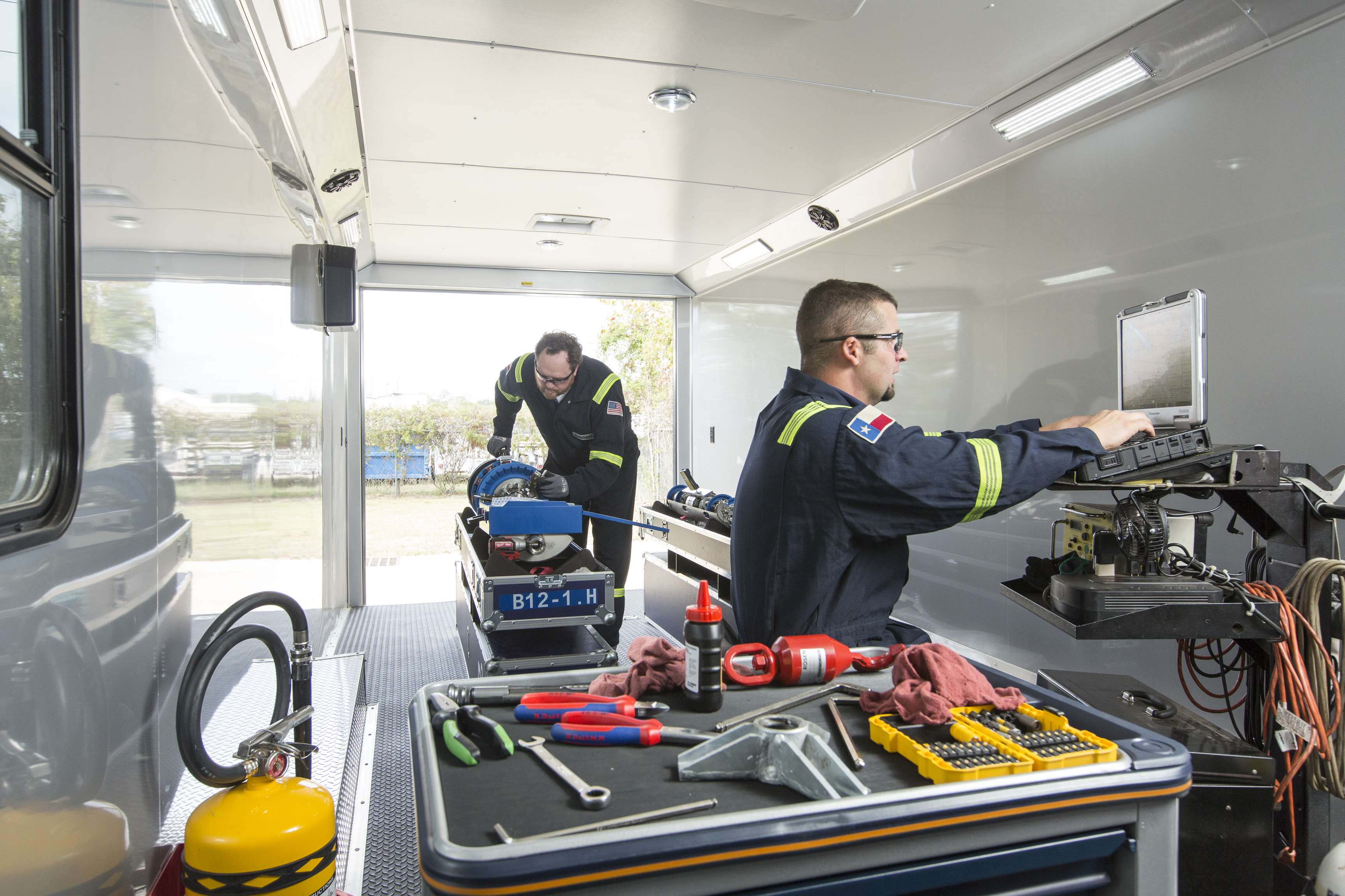 Two field technicians working in the mobile diagnostic unit the left one is working on a tool the other at a laptop.