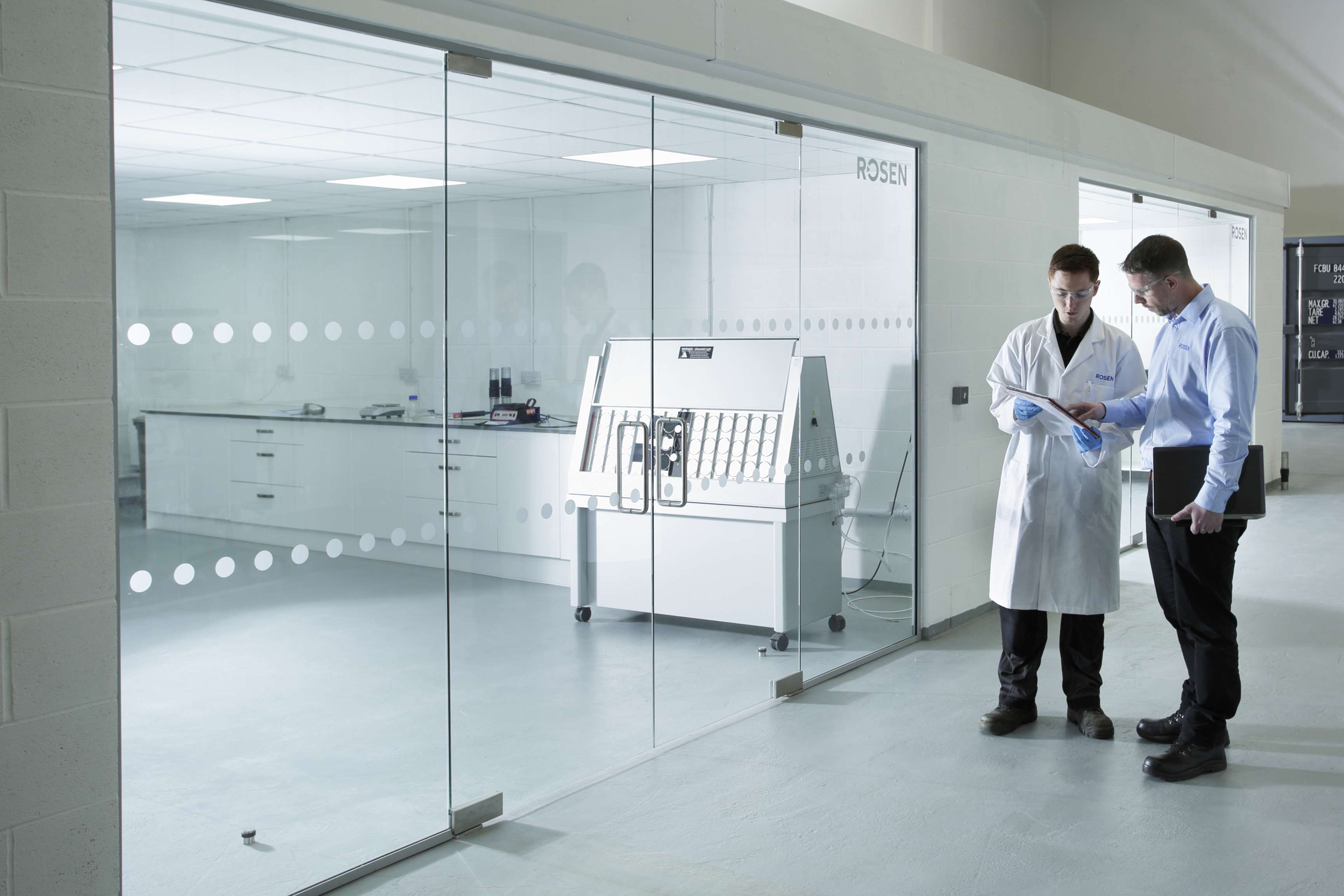 Two employees wearing protective goggles stand in a corridor of a laboratory and look together at a clipboard.