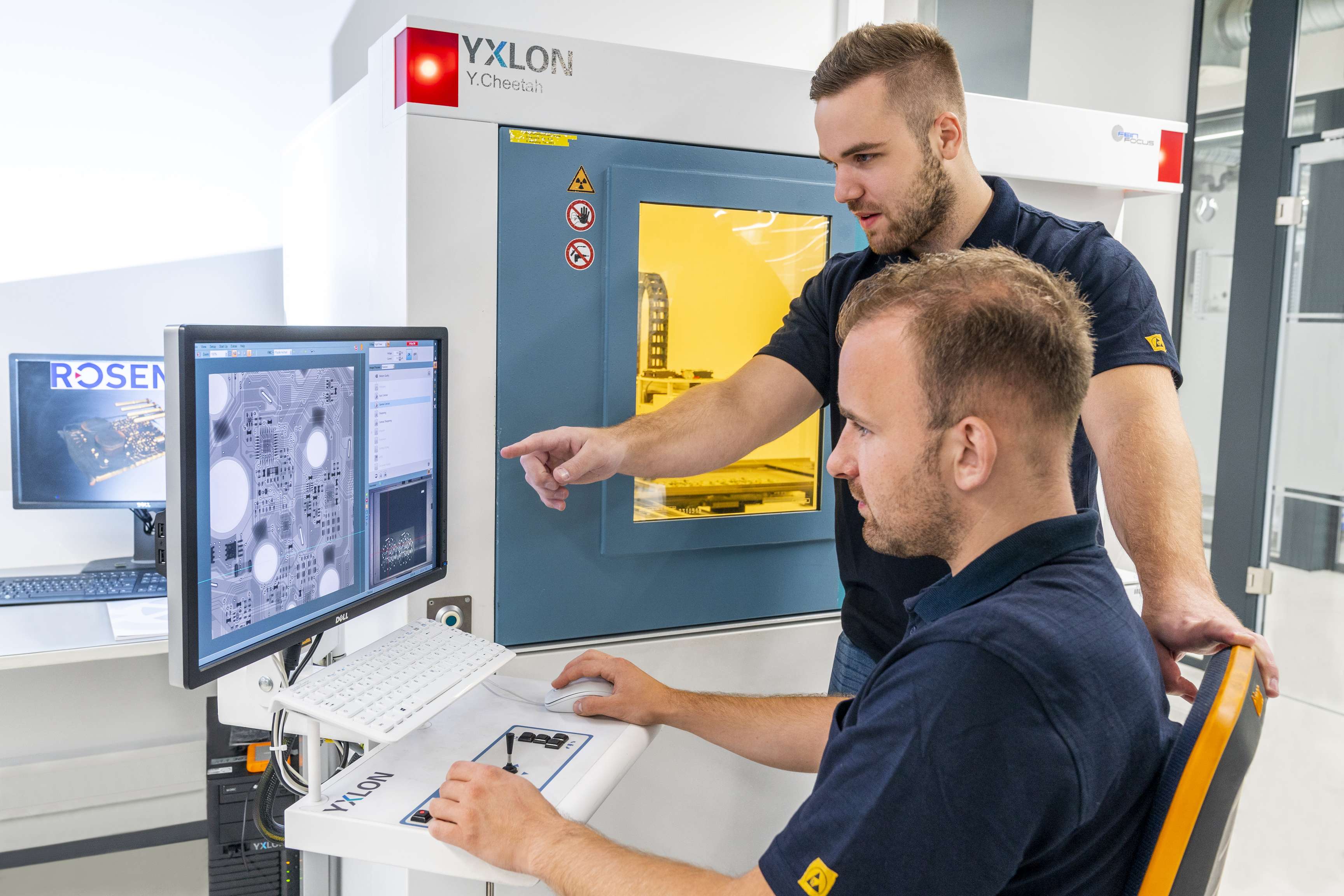 Ein Kollege sitzt an seinem Schreibtisch, ein Kollege steht neben ihm und zeigt Daten auf dem Bildschirm.