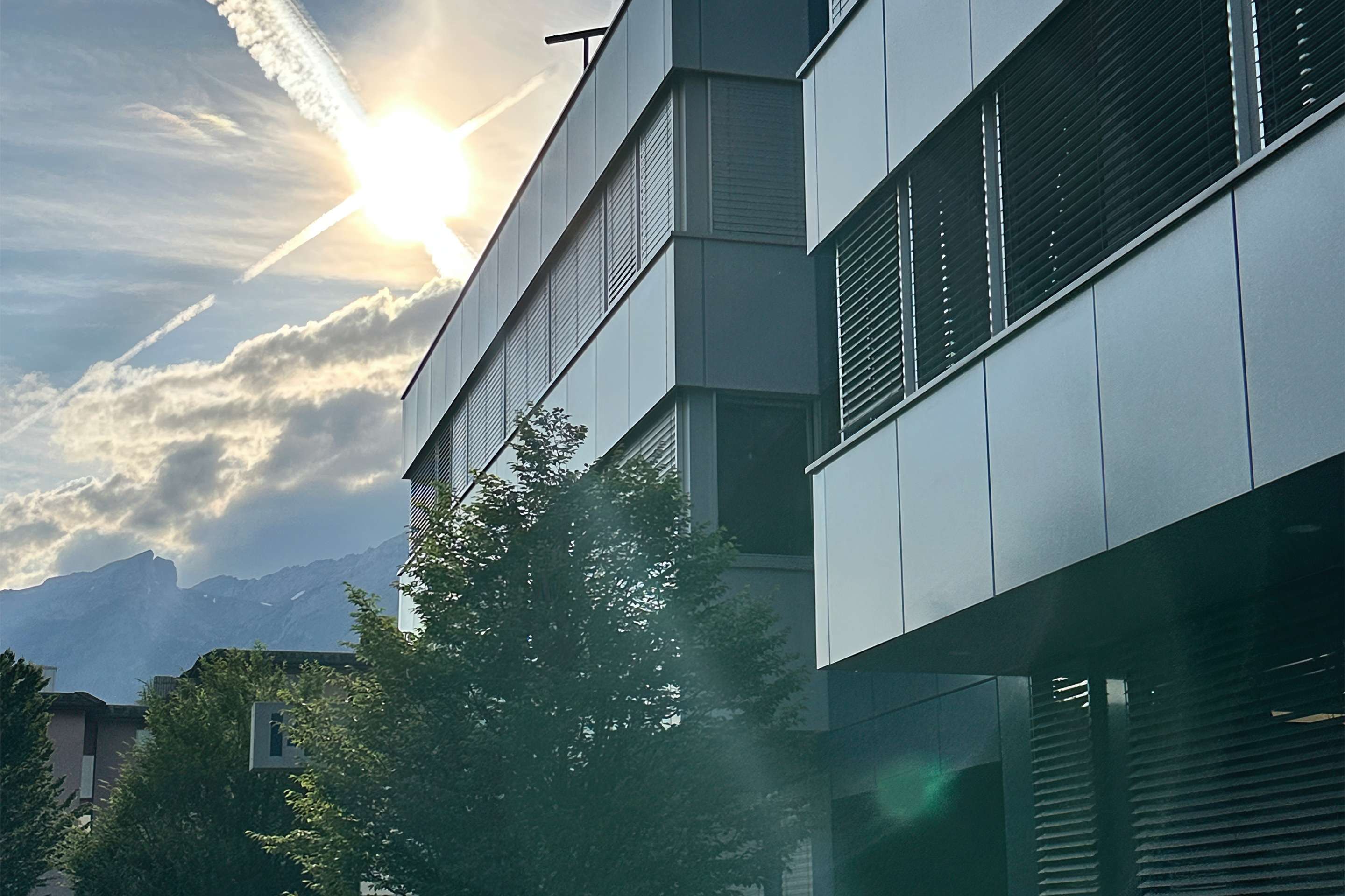 The photo shows the ROSEN Group headquarters in Switzerland, with its metal facade, backlit by the sun and surrounded by trees, with a mountain landscape in the background.