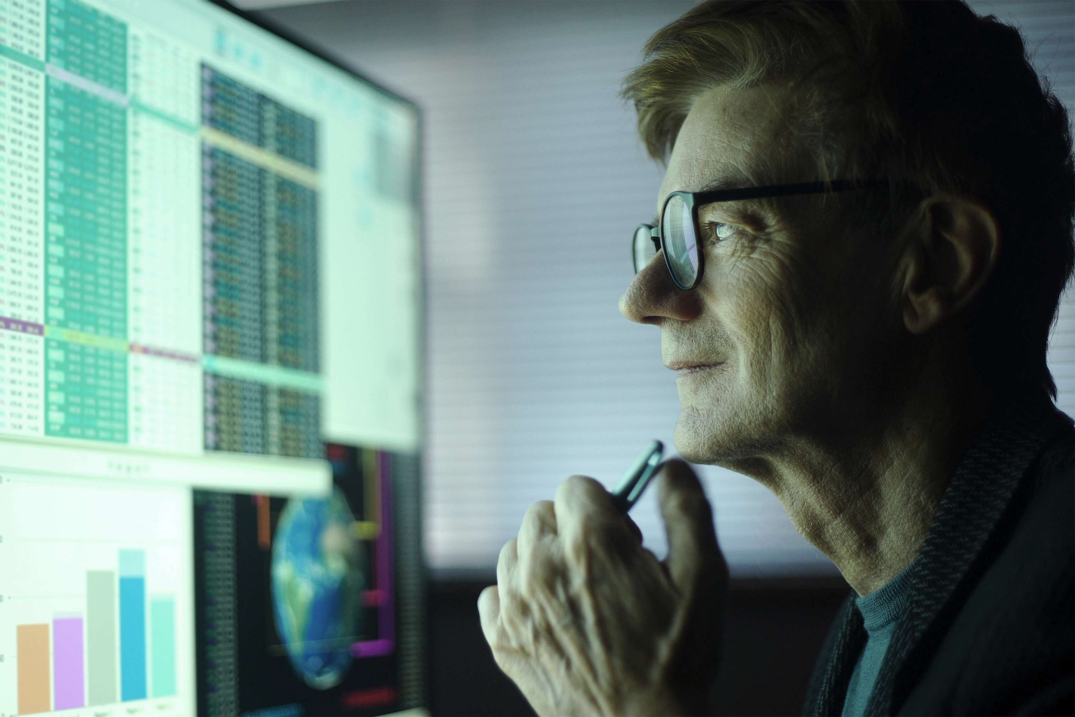 Man with glasses in front of a screen that shows charts and data.