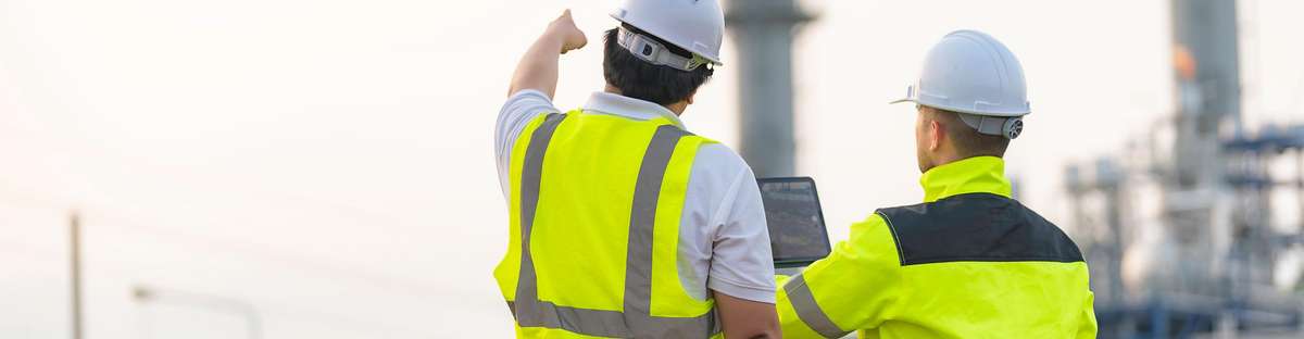 Two experts from behind in front of a refinery. One is pointing at something in the distance, the other is holding a laptop.