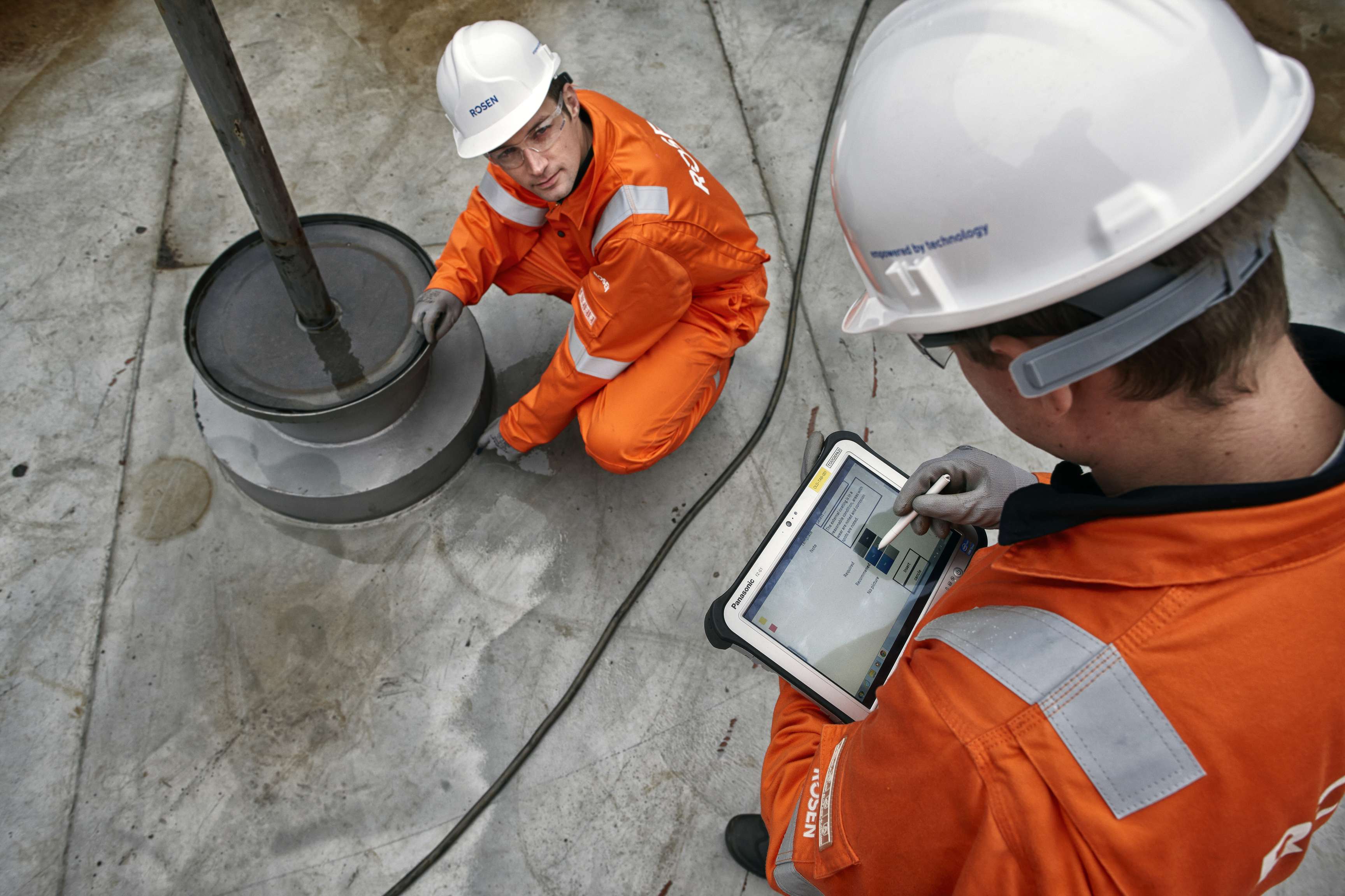 Two ROSEN experts in a tank inspecting the tank bottom. One kneels and looks up at his colleague, who is looking at his tablet in his hand.