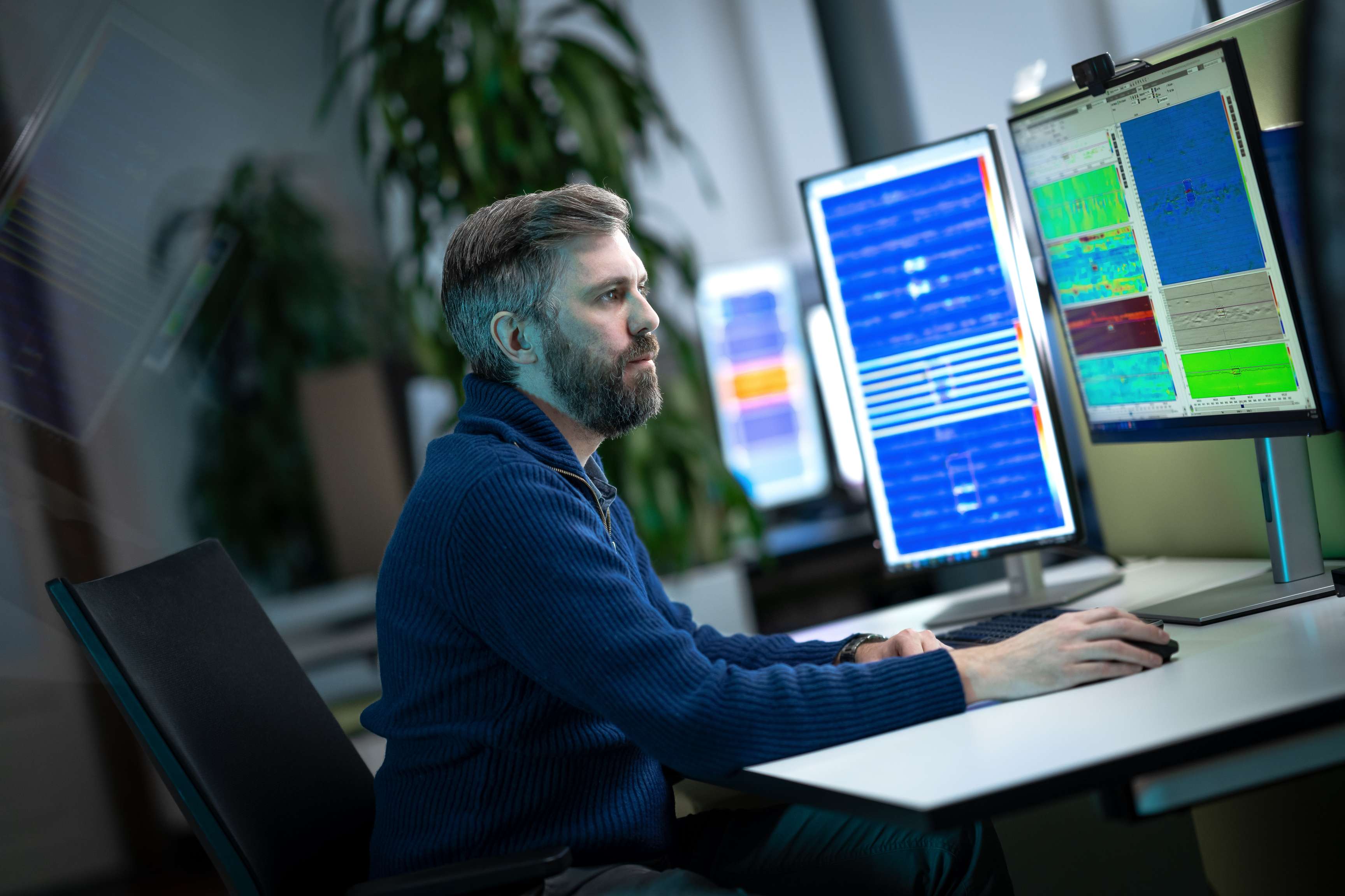 A man is sitting at a desk in an office. On the desk are screens that he is looking at and working on with various data and records.