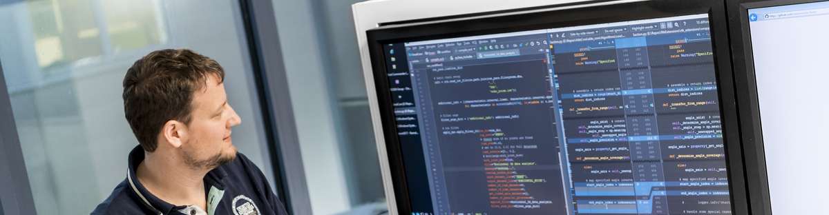 Ein Kollege aus der Abteilung für Softwareentwicklung steht an einem Stehtisch mit seinem Laptop und schaut auf den Präsentationsbildschirm.