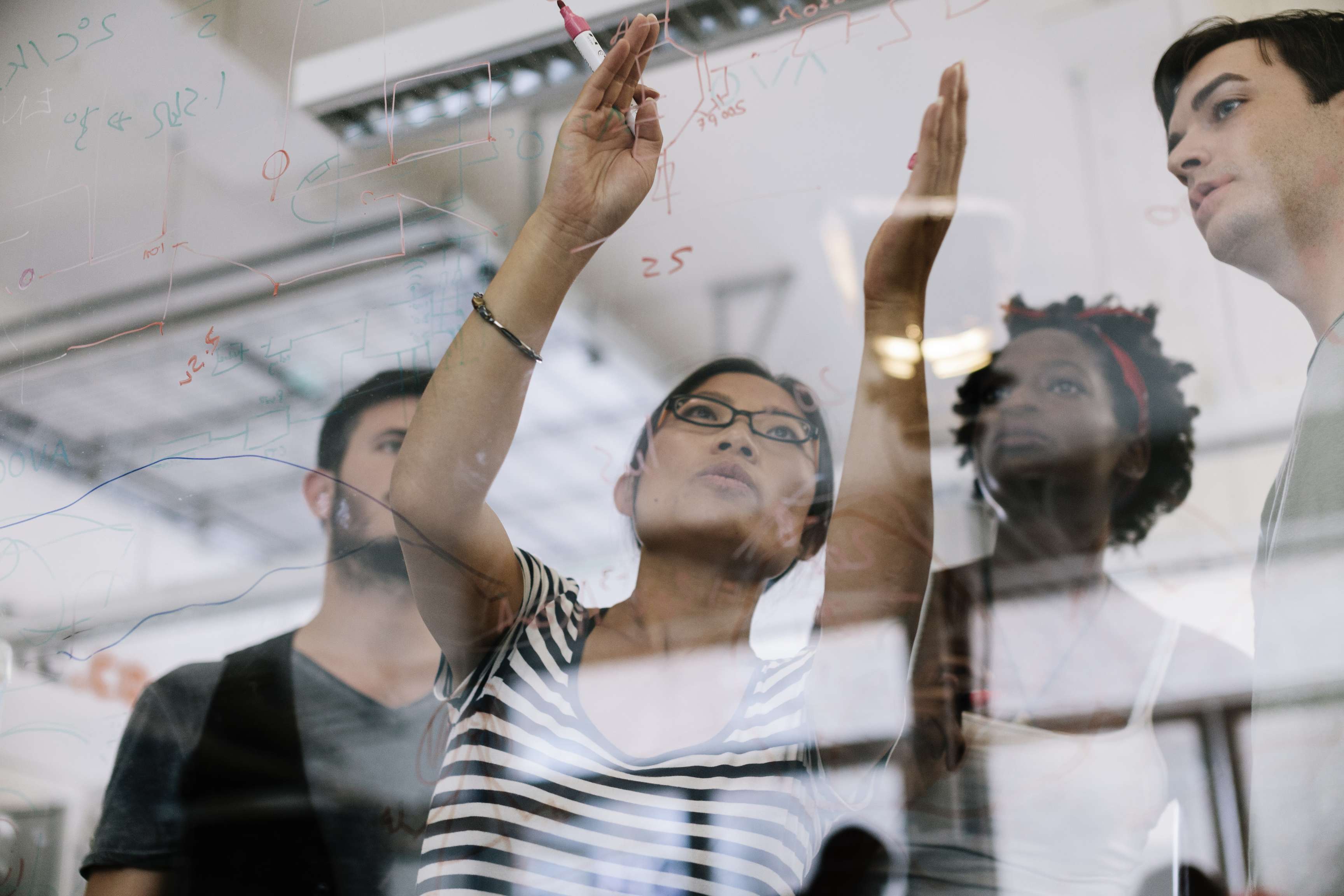 A woman leads a discussion about a project with three other people by pointing to a mathematical equation on a glass wall.