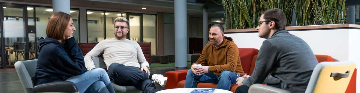 Four people are sitting on colorful armchairs around a small round table in a modern, open-concept space at the Lingen site, engaged in conversation. The room features glass walls, plants, and an open-concept design. The image is from the “IT Software Data Science” campaign from 2024 and depicts a conversation taking place in a work environment.