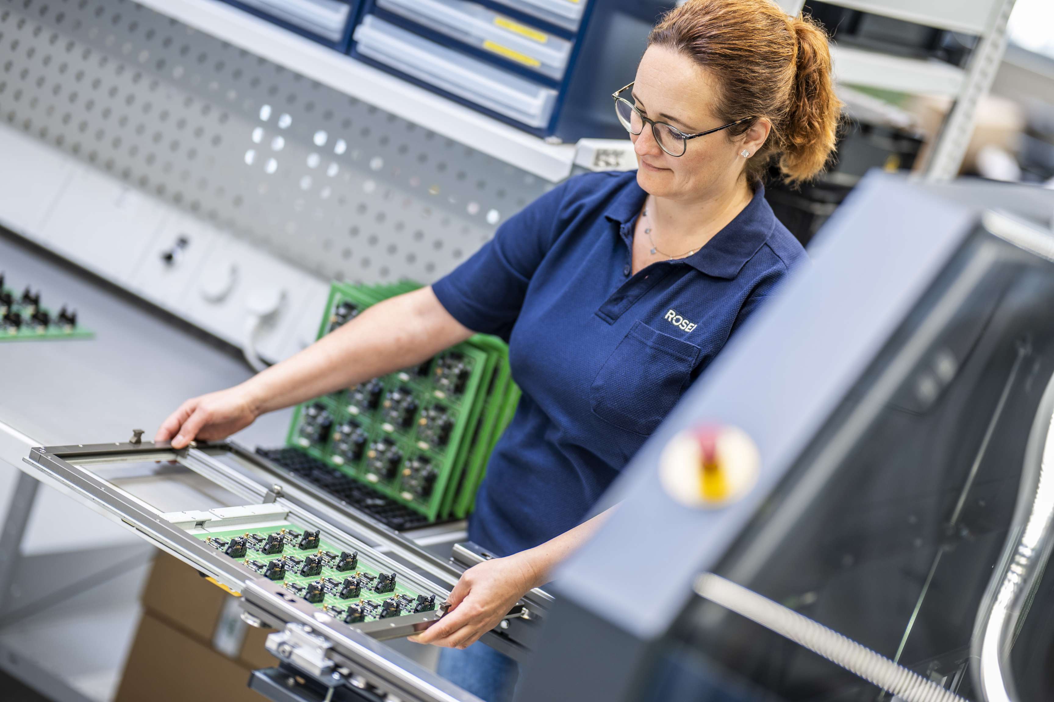 Eine Frau steht in der Elektronikfertigung und hält eine Platine in der Hand
