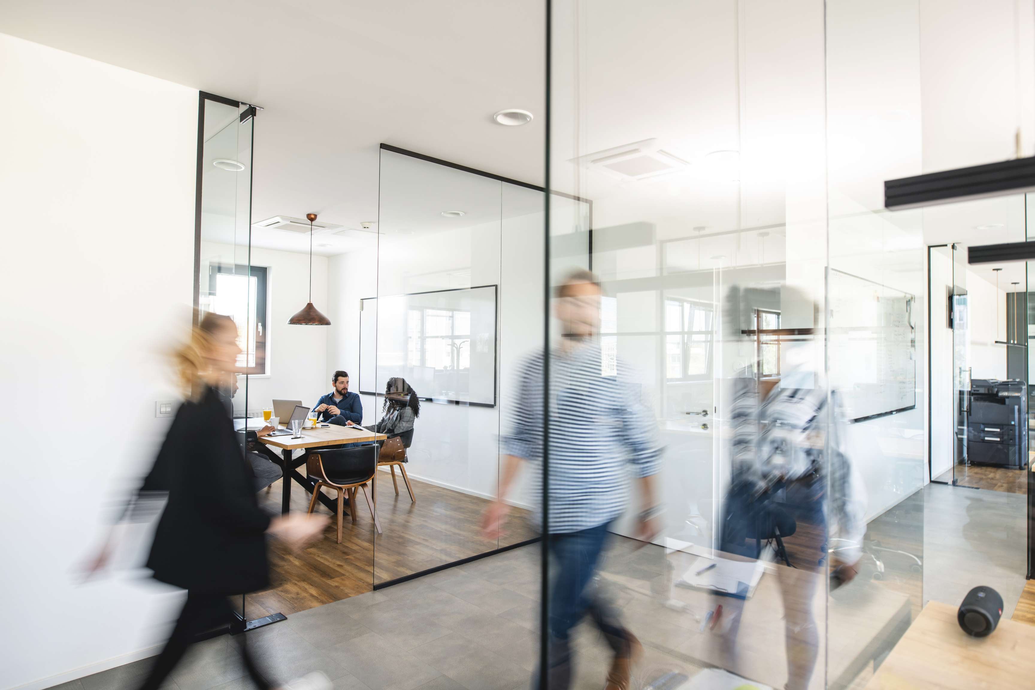 Three people are sitting in a meeting room made of glass and are talking while some people are walking by in front of the room.