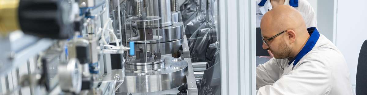 Employee works in a booth in a laboratory while his colleague stands next to him and watches.