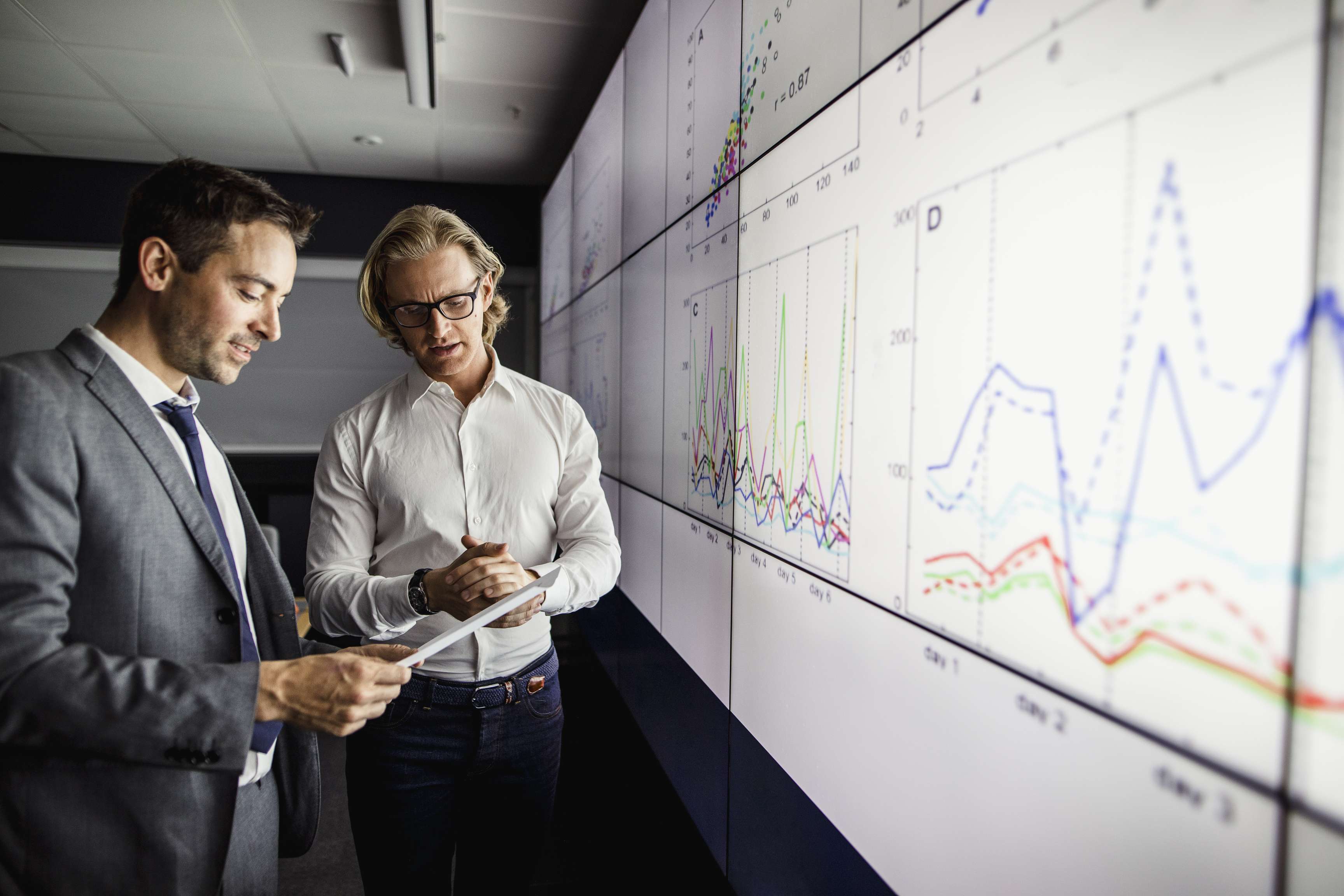 Group of business people in a dark room standing in front of a large data screen with information.