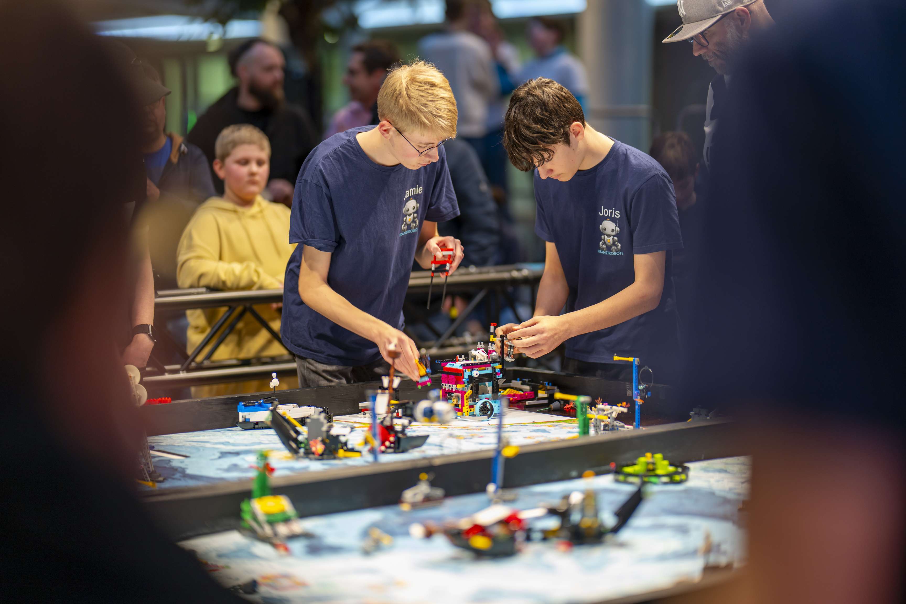 Two boys are working on their LEGO robot at a table. Onlookers are watching them.