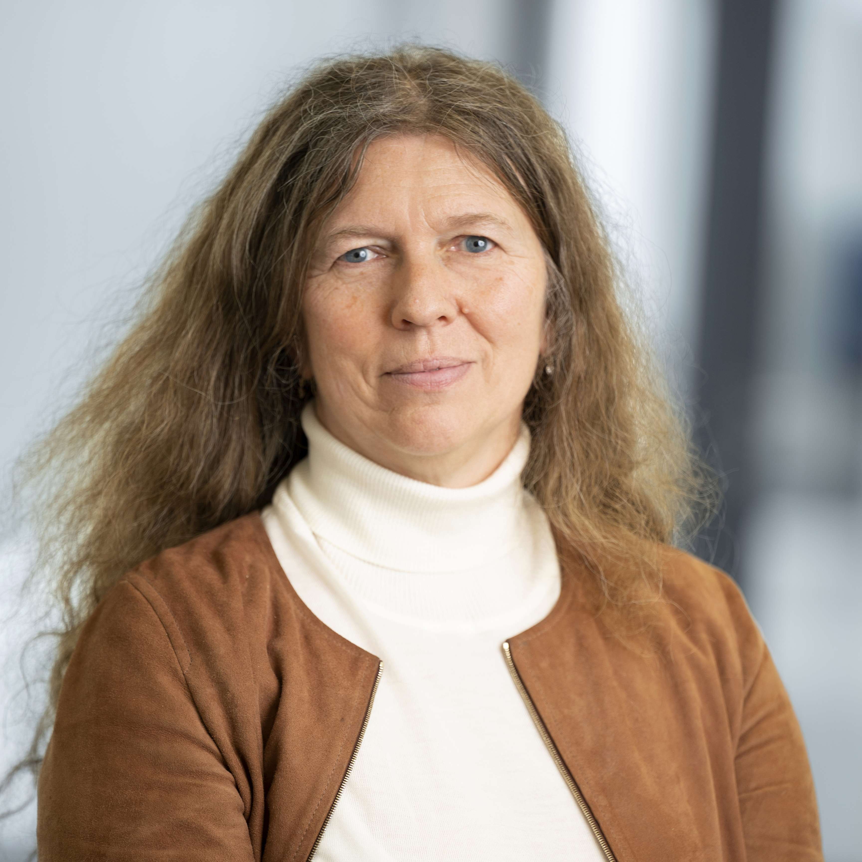 Woman with brown long hair smiles at camera wearing brown jacket over white turtleneck sweater.