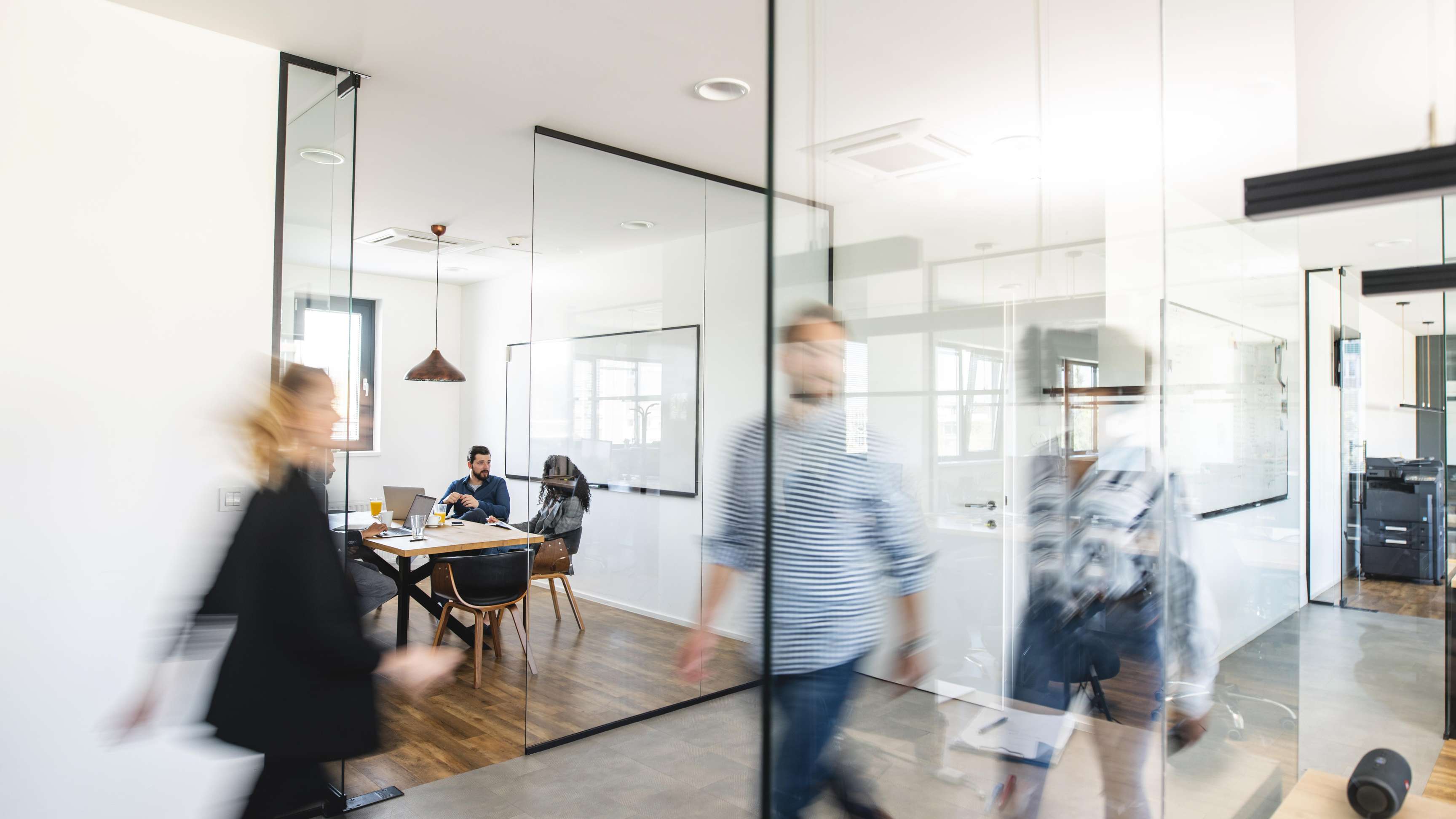 Three people are sitting in a meeting room made of glass and are talking while some people are walking by in front of the room.