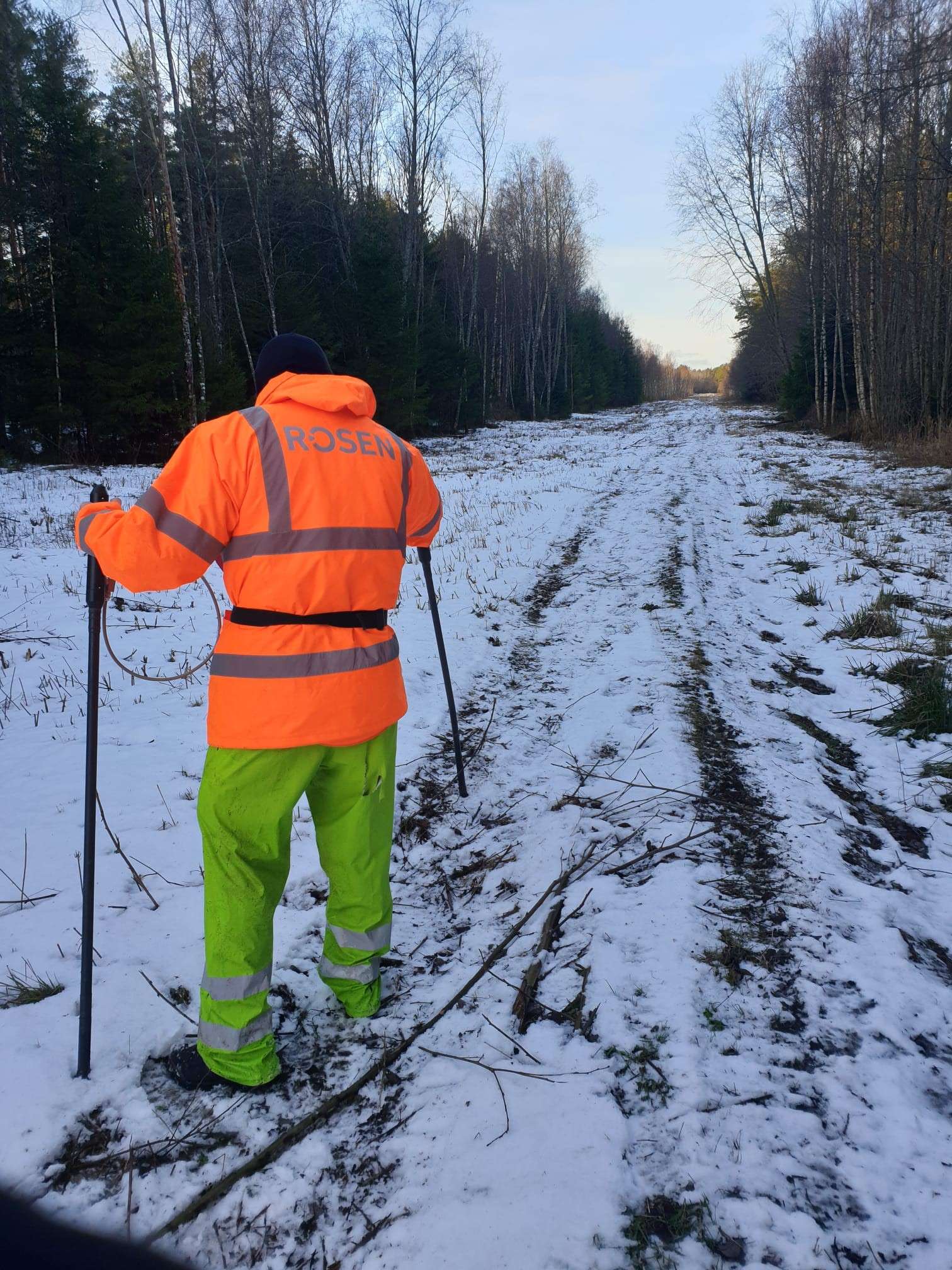 Un empleado de ROSEN con chaqueta naranja neón con el logotipo de ROSEN en la espalda y pantalones amarillos se encuentra en un paisaje nevado con un bosque a cada lado y marcadores en las manos.