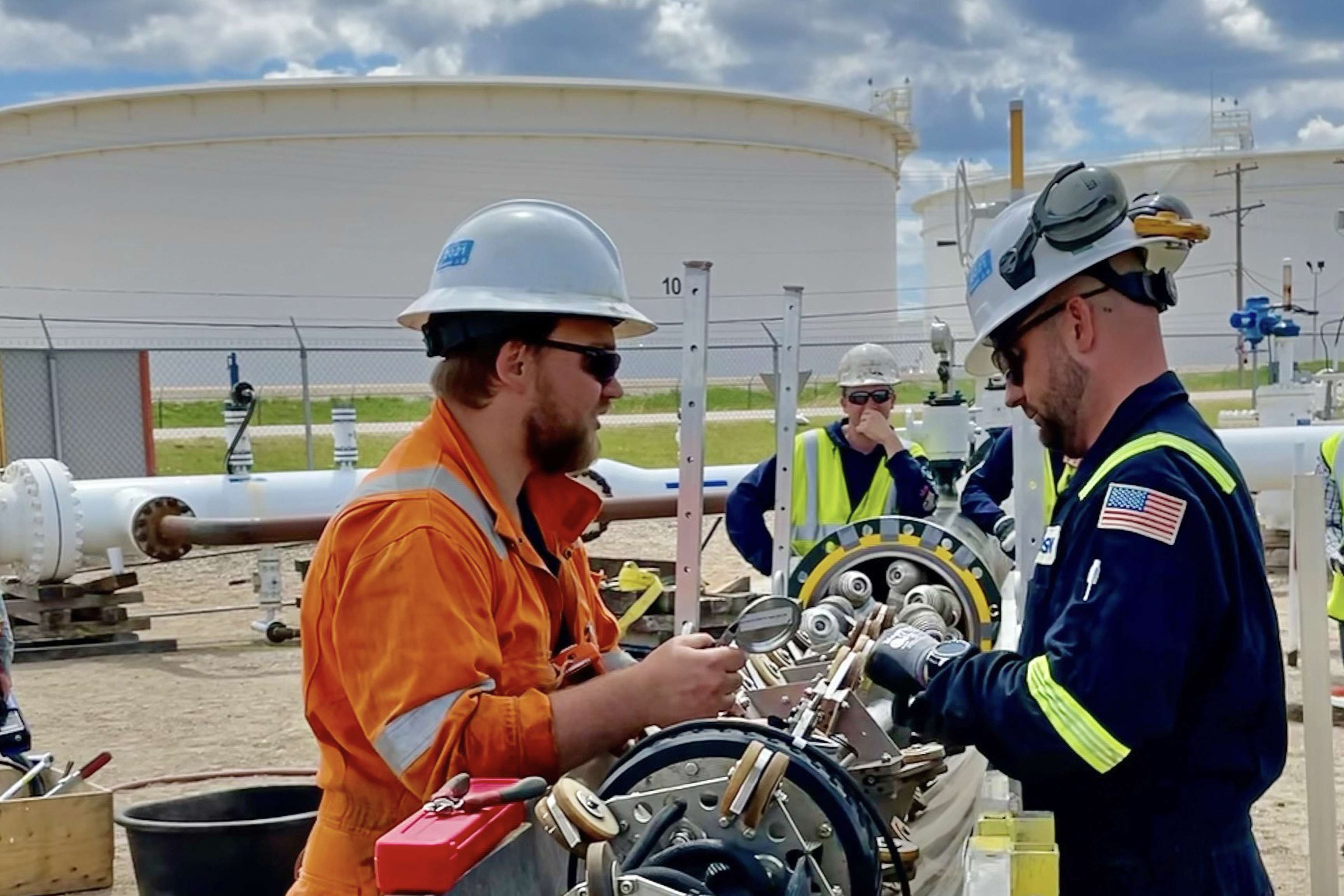 Tethered tool before it goes into a pipeline, with a ROSEN experts in orange and blue overalls inspecting it, with more white Pipelines and tanks in the background.