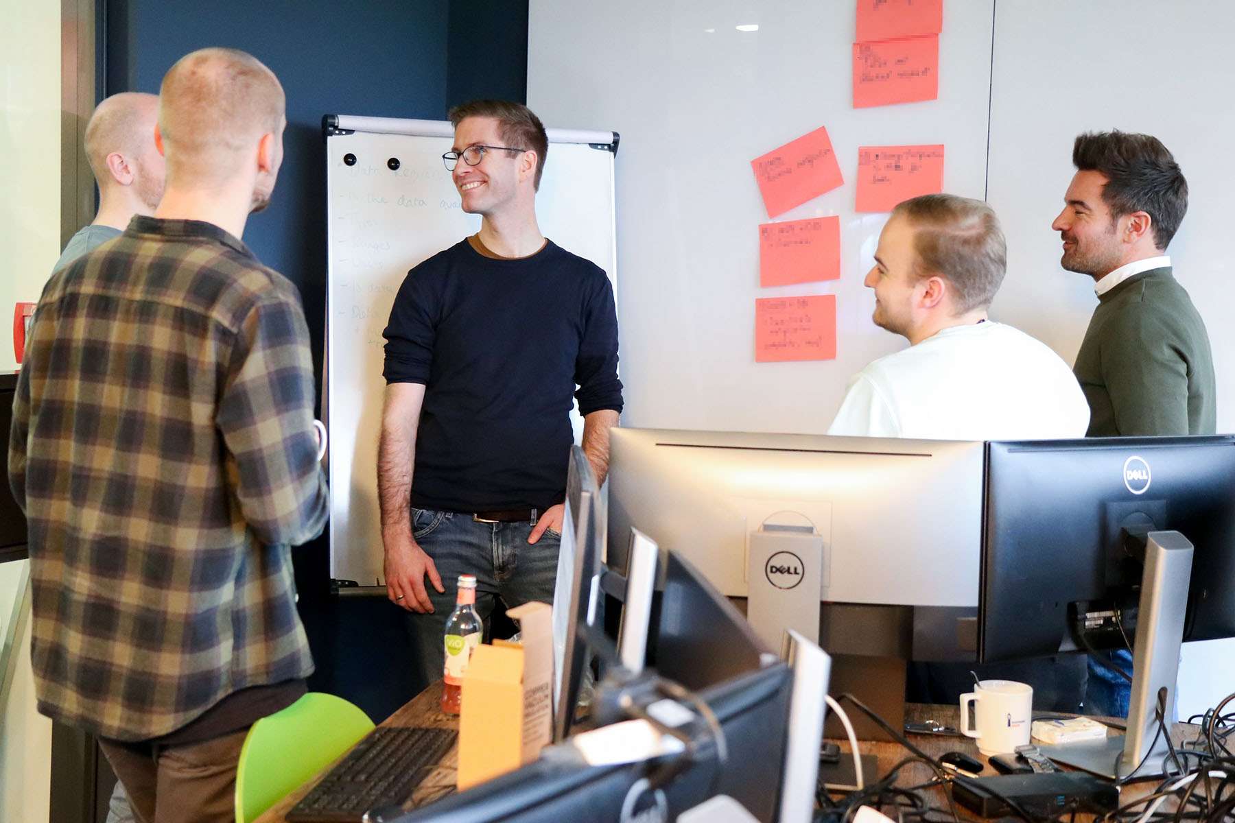 A group of men are standing in front of a flip chart and computer monitors, talking.