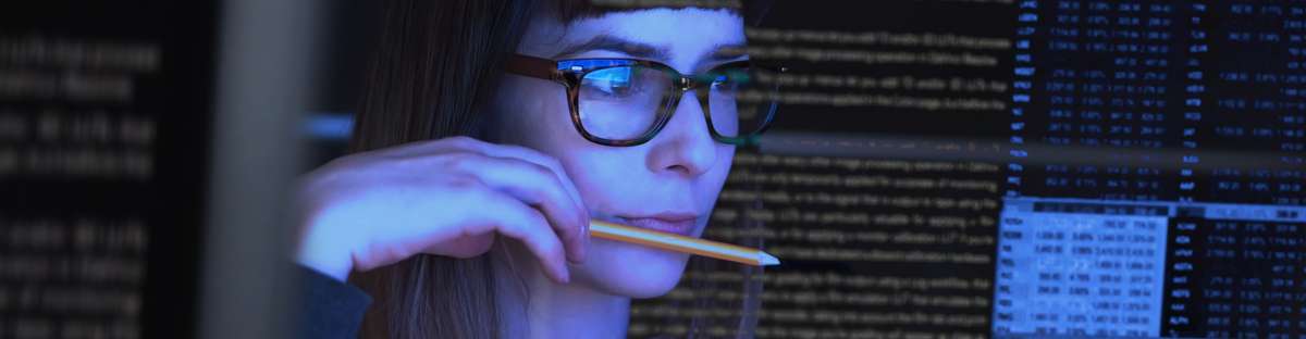 Woman wearing glasses and holding a pencil in her hand and putting it to her lips while looking at a screen with data sets.