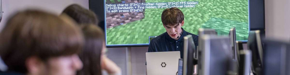 Schüler arbeiten an Computern im Klassenzimmer, während ein Referent auf einer großen Leinwand eine digitale Anwendung erklärt.