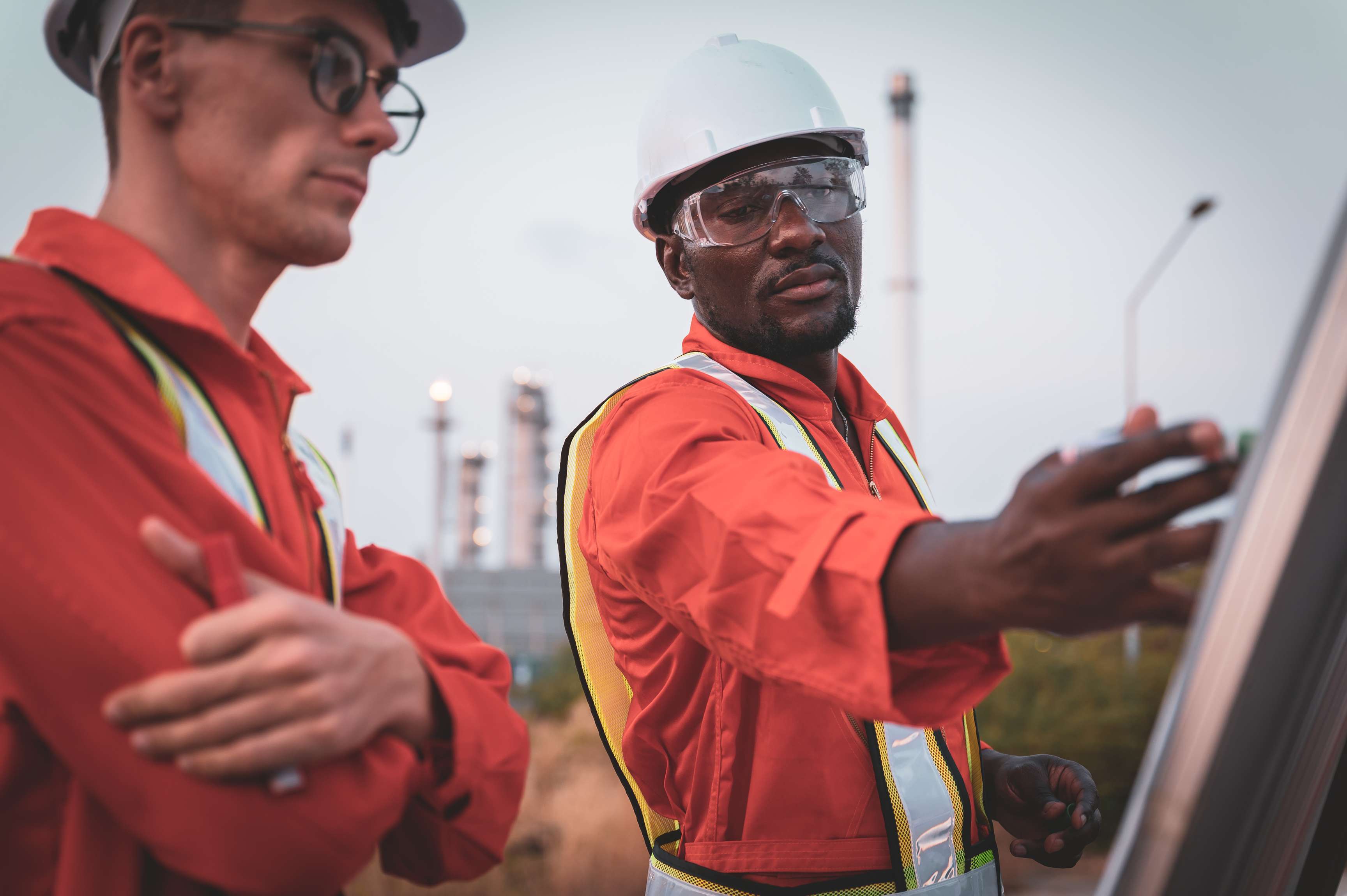 Two men in orange coveralls standing in front of a flip chart looking at it with a refinery in the background.