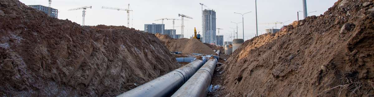 Two pipelines in the ground with backfilled earth on both sides, city with cranes in the background.
