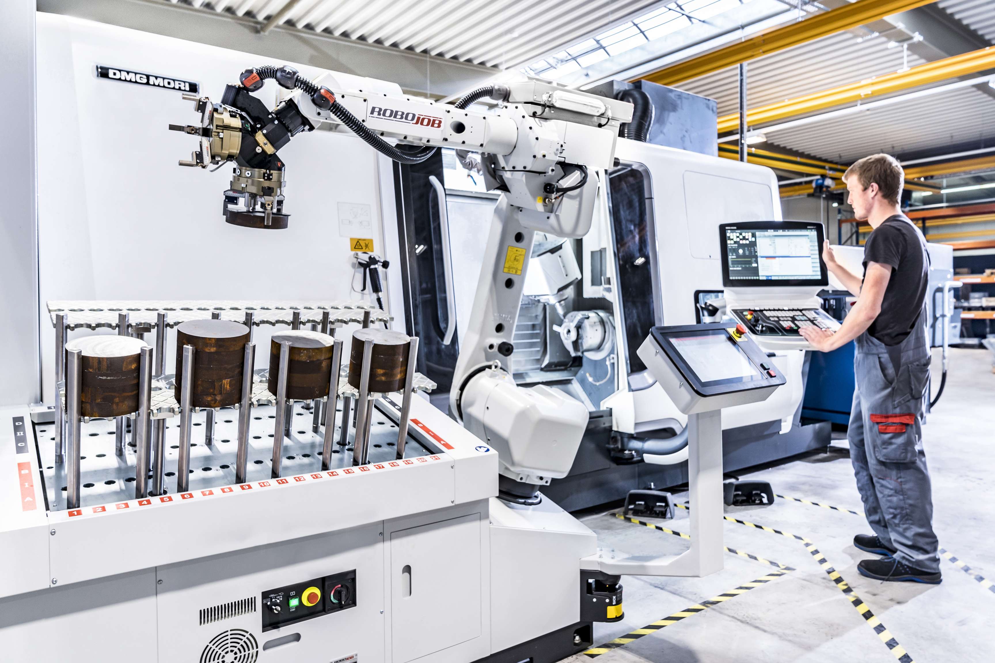 Ein ROSEN Mitarbeiter steht an einer hochkomplexen Maschine und steuert sie mithilfe des außen angebrachten Monitors. Links im Bild sieht man fertig hergestellte Bauteile, die von einem Greifarm gestapelt werden.