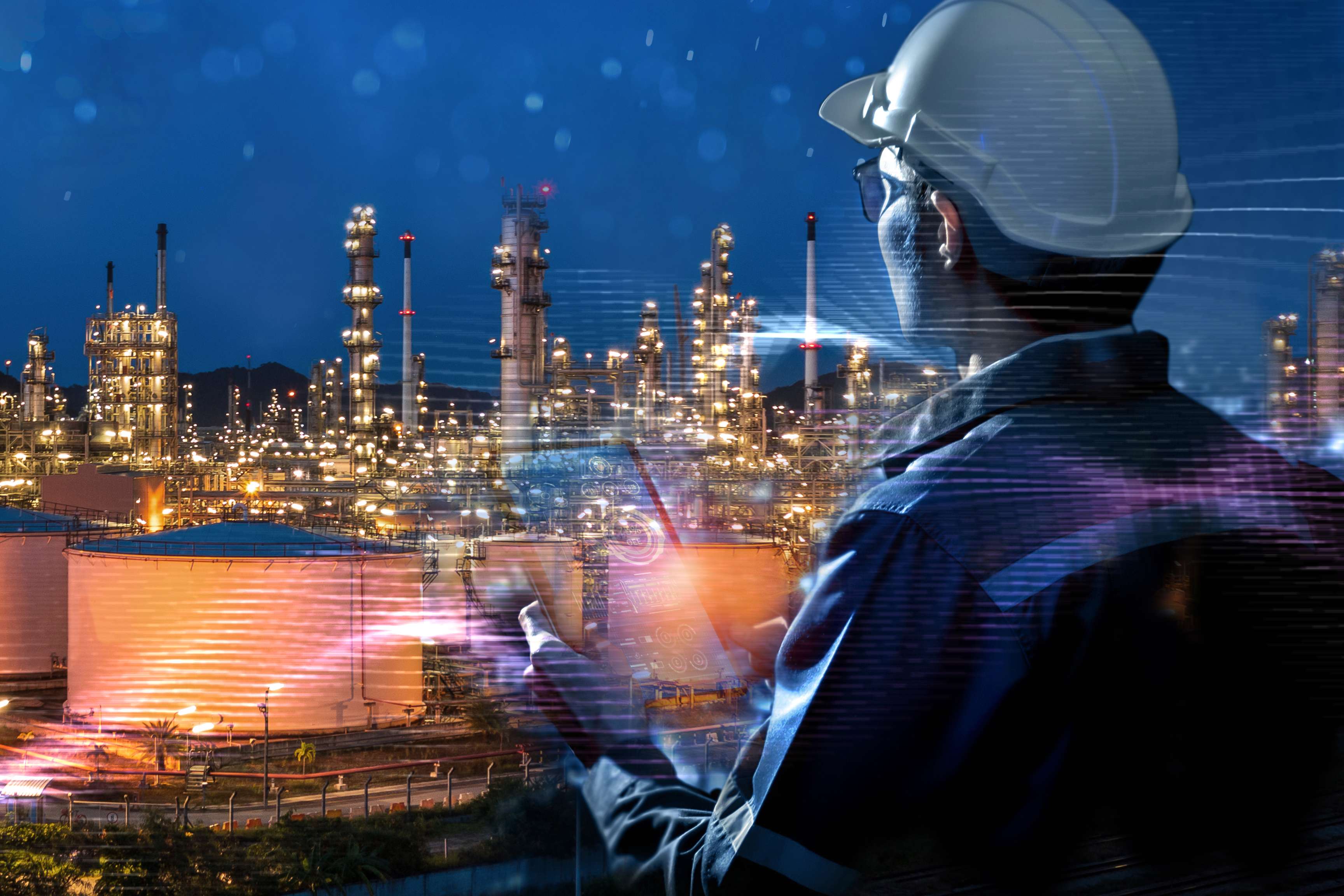 Refinery site in the dark with many lights shining on it and a man in the foreground looking towards the refinery site, wearing a safety helmet and holding a tablet in his hand.