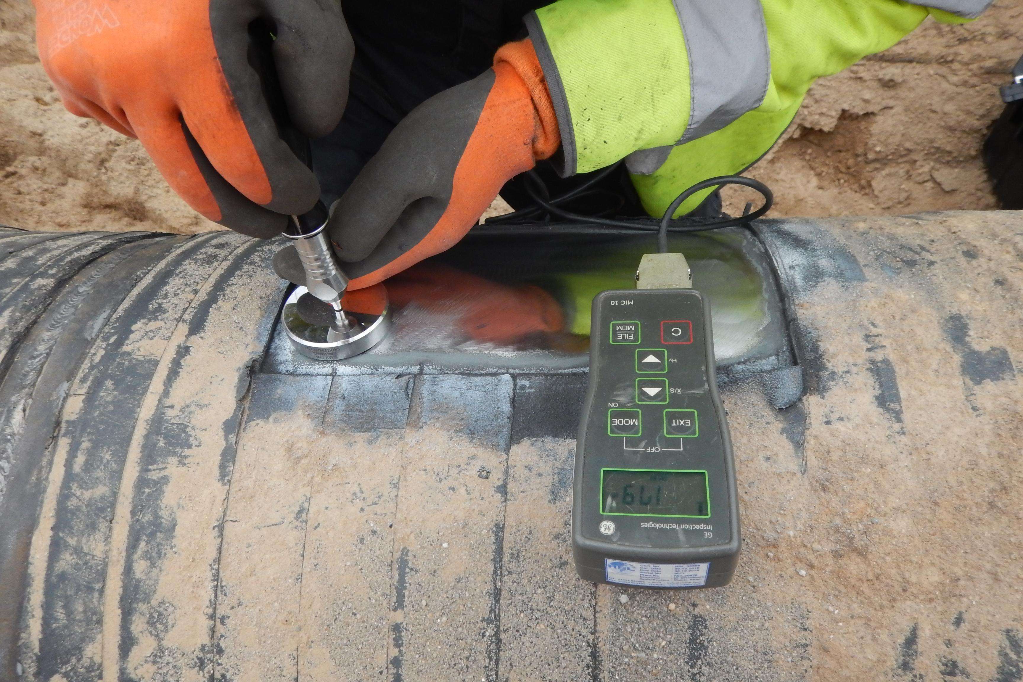 Close-up of a dirty pipeline with a measuring device on it, while hands in gloves work on it with a silvery part.