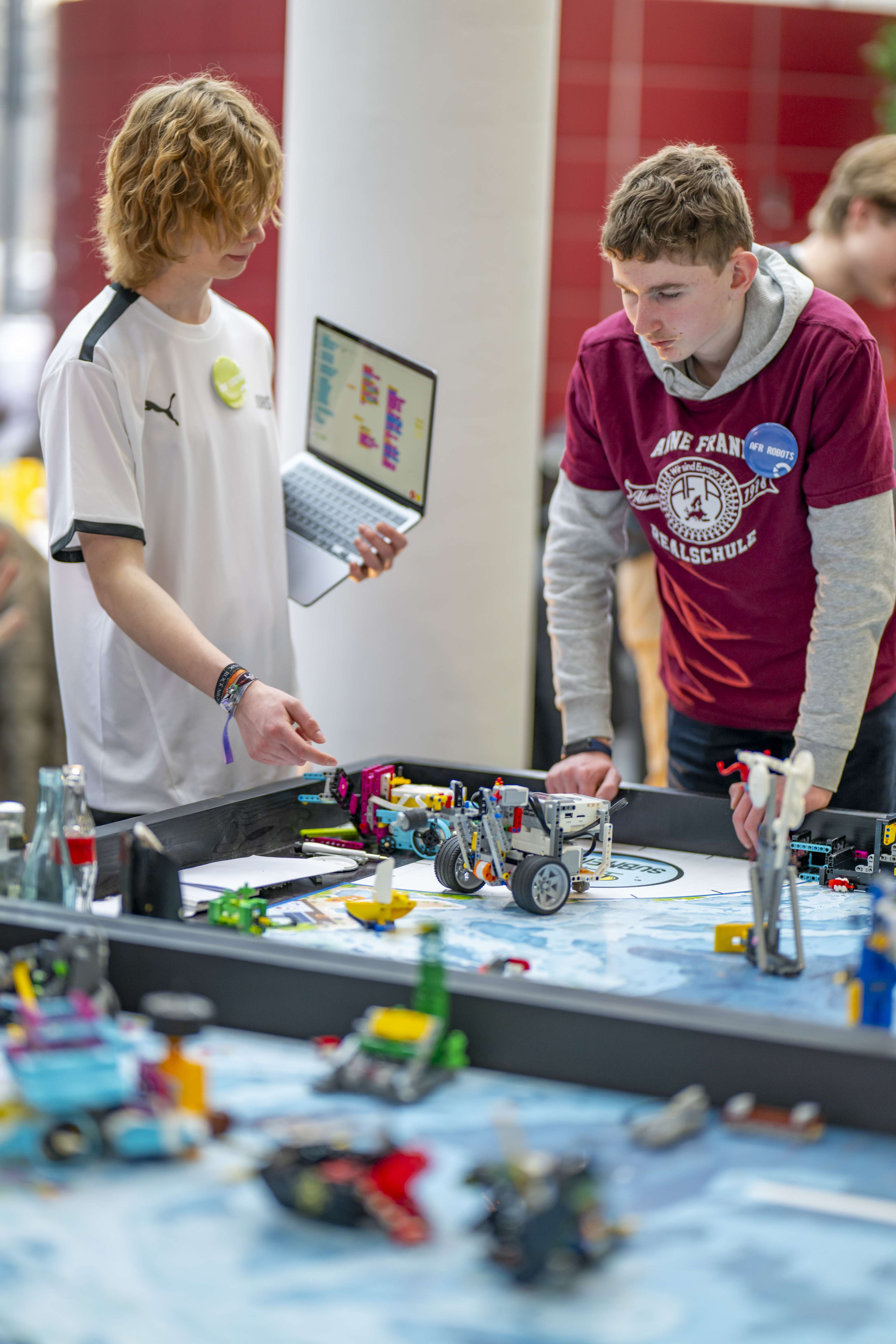 Two boys are standing at a table with LEGO robots and talking.